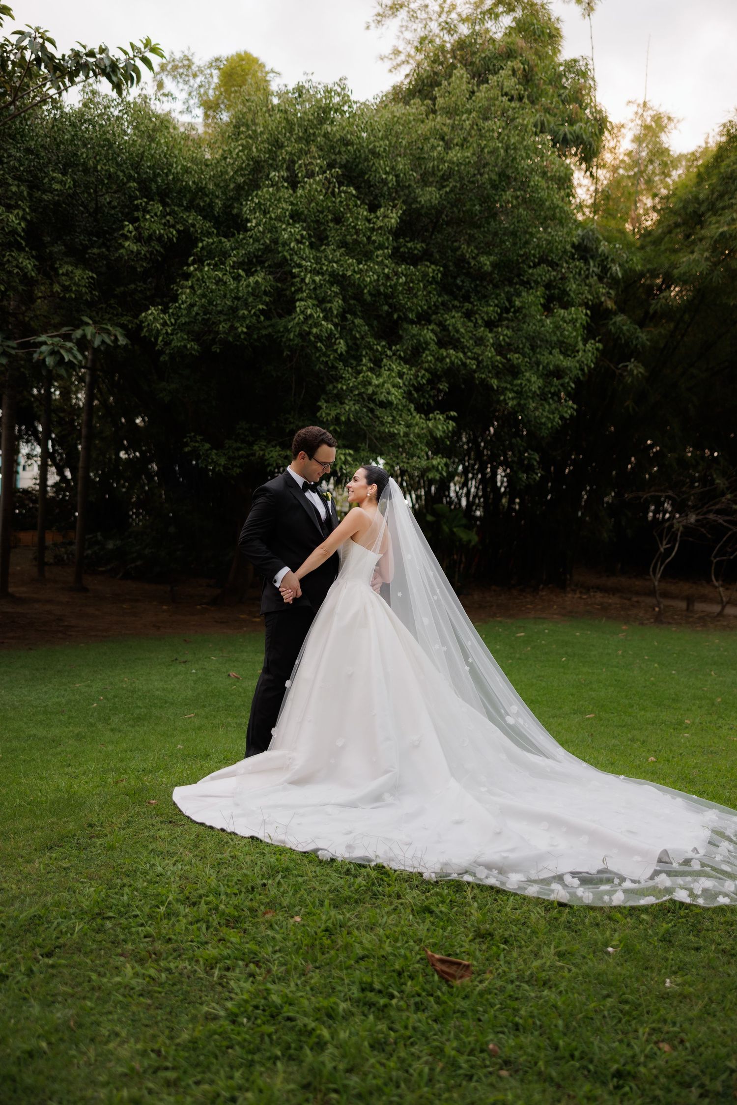 A bride and groom pose together on a grassy lawn with a flowing white wedding dress trailing behind them.