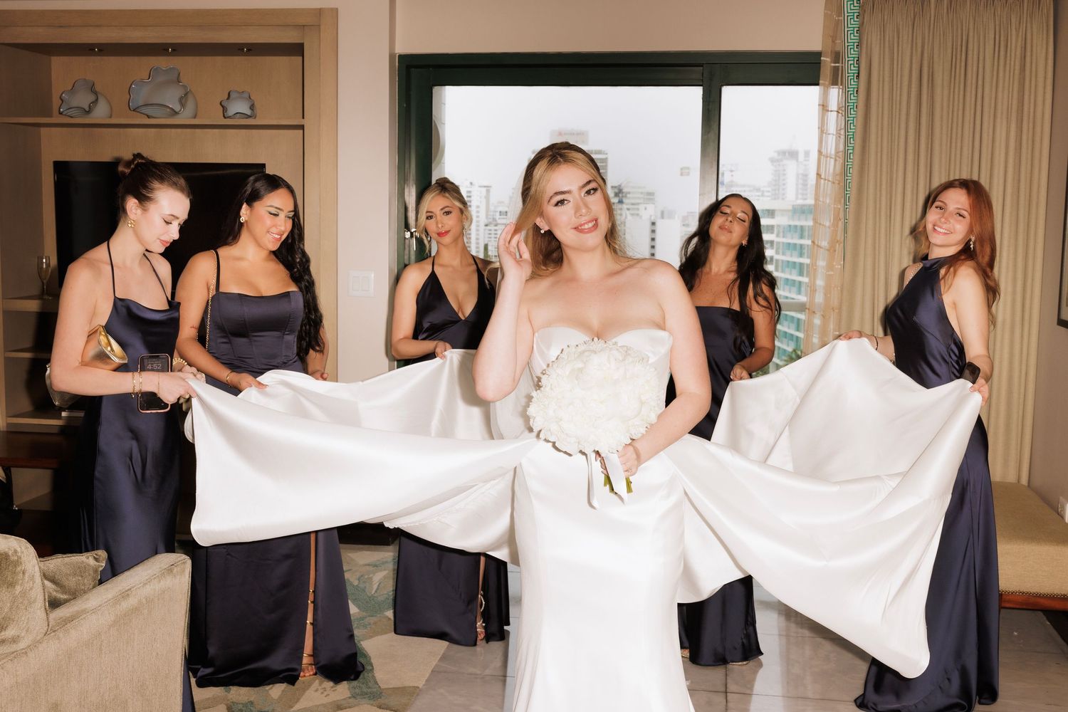 Bridesmaids in navy dresses help arrange the bride's gown in an elegant hotel room setting.
