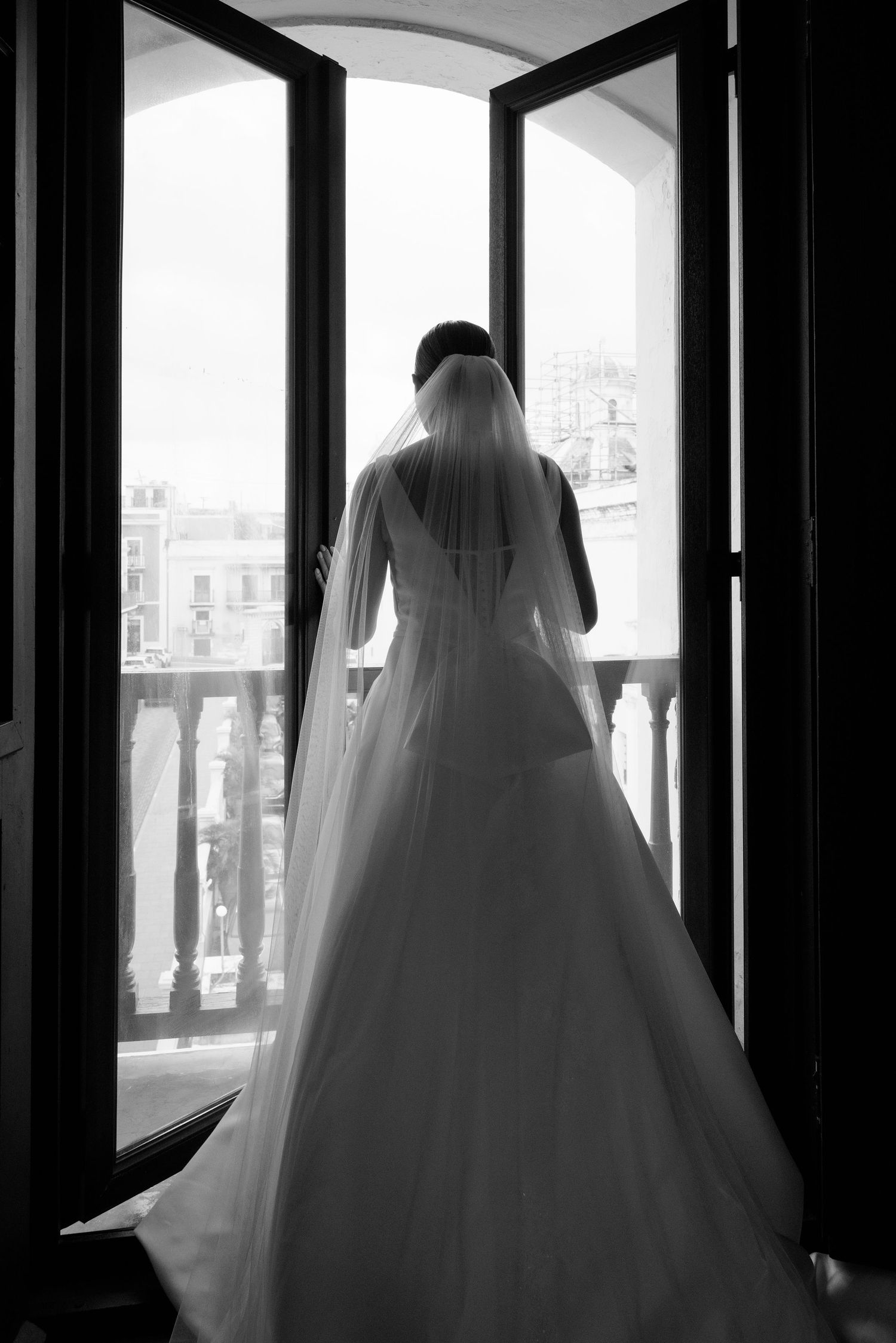 Dramatic black and white silhouette of a bride in a ballgown wedding dress framed by large windows.
