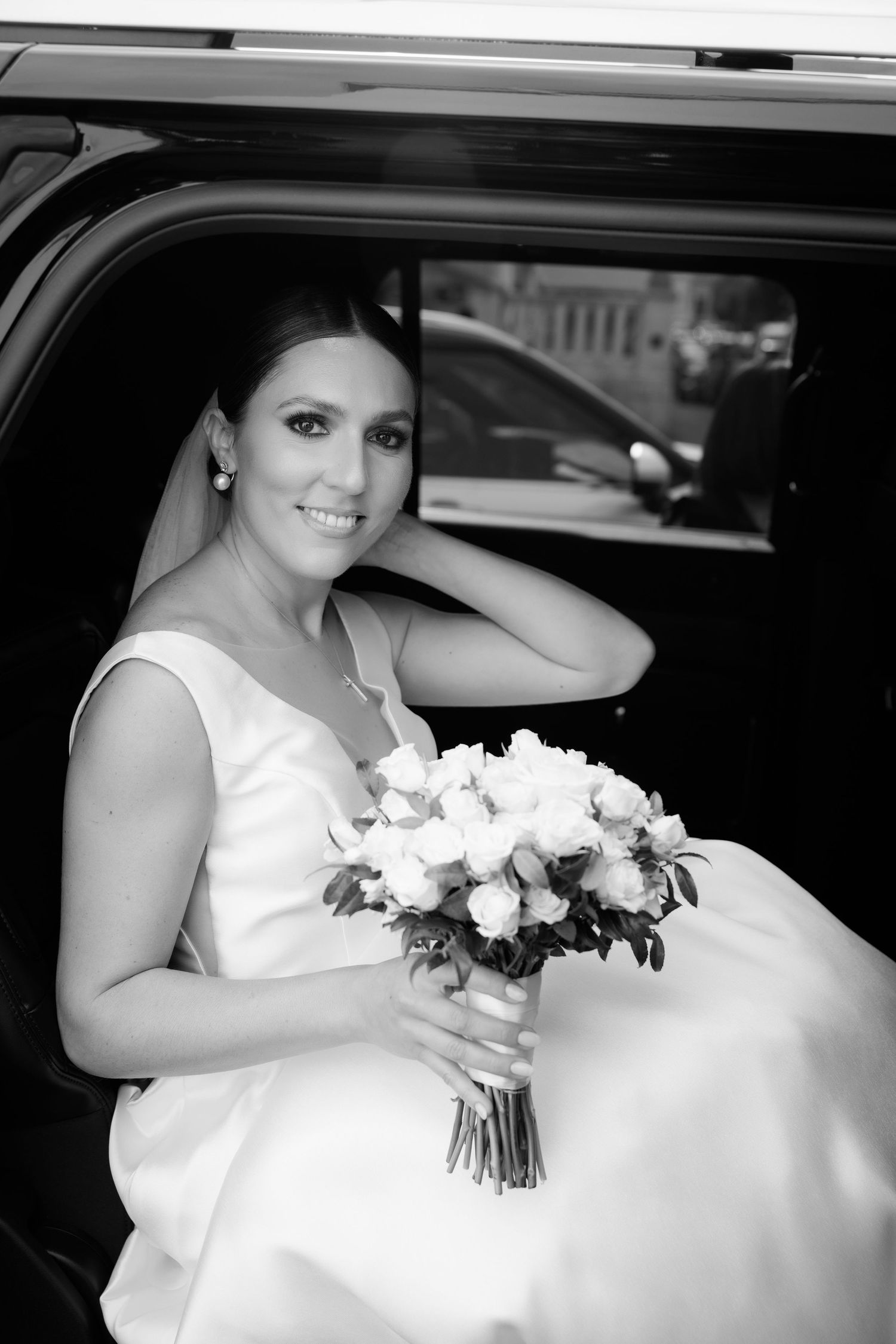 A bride in a white wedding dress holds a bouquet while sitting in a car on her wedding day.