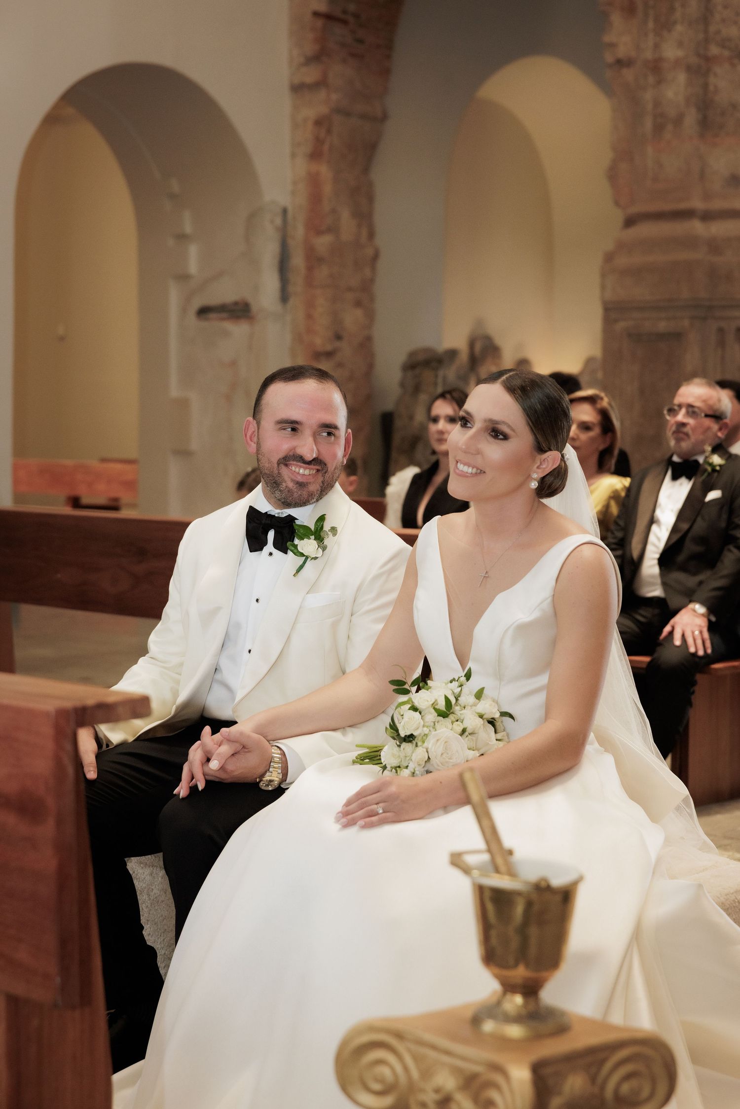 A wedding couple sits together in an ornate church with stone archways and columns.
