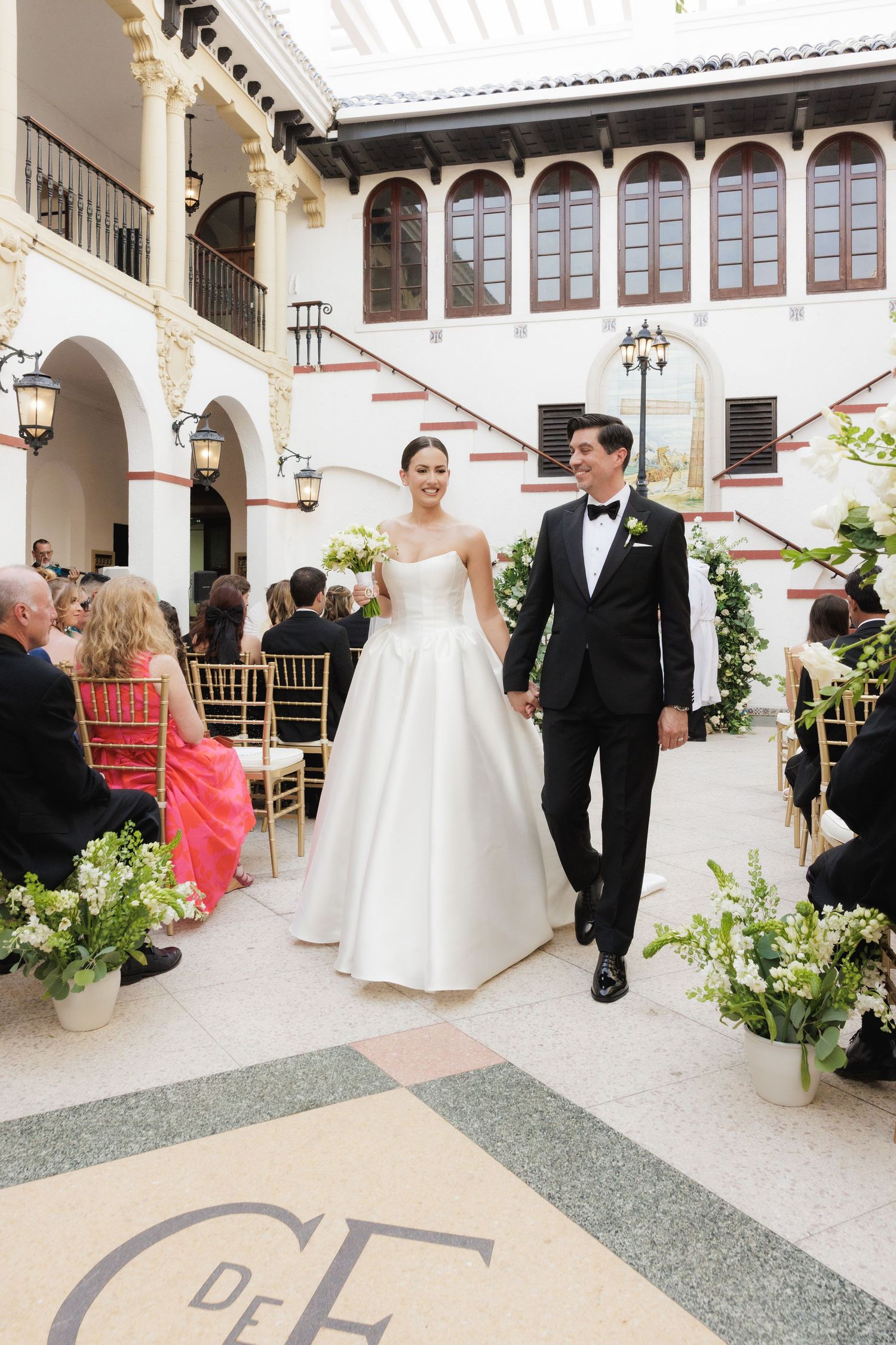 Wedding couple walks through elegant Spanish colonial style courtyard with white arches and decorative lighting.