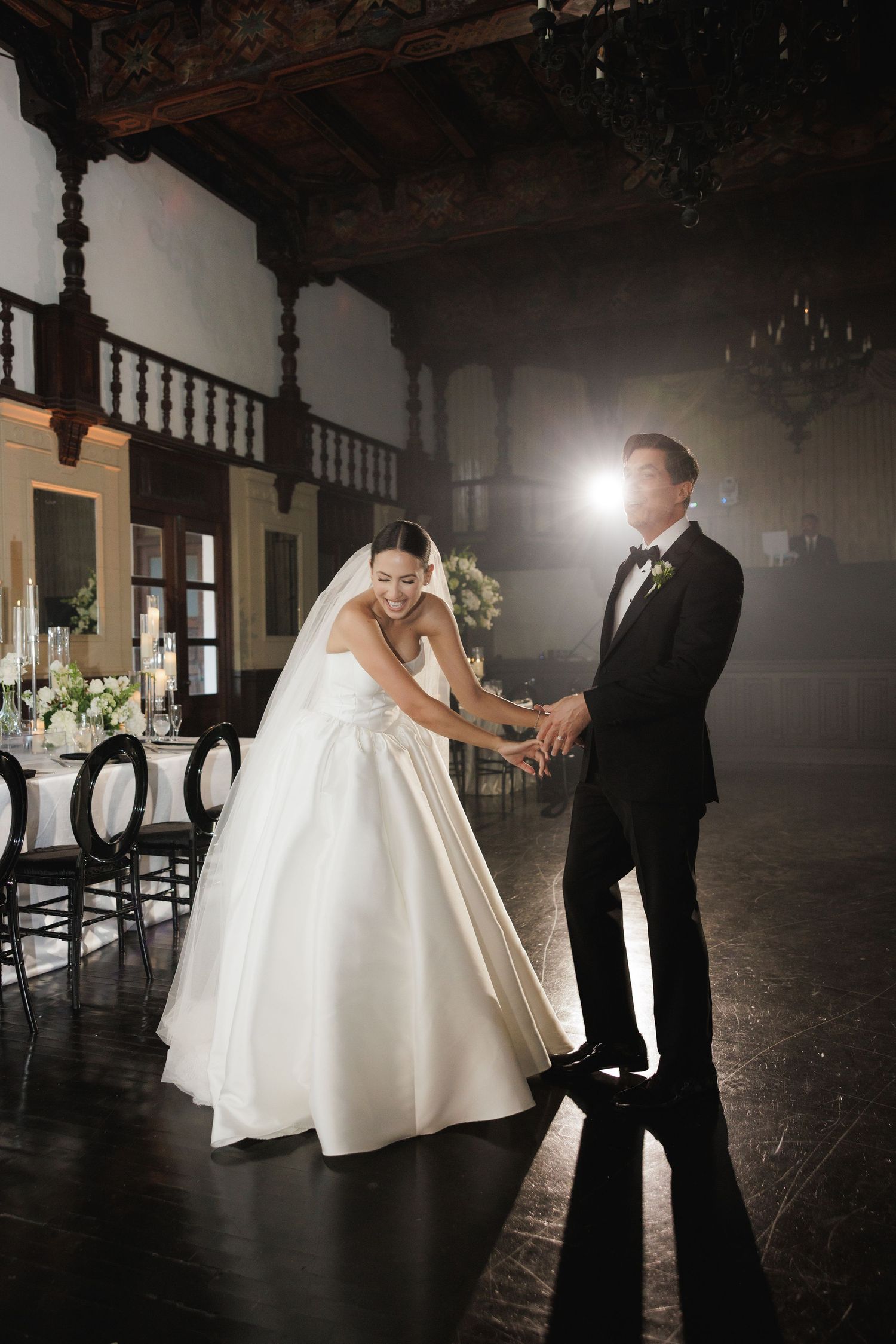 A couple shares a romantic dance moment in a dimly lit ballroom with ornate wooden details.