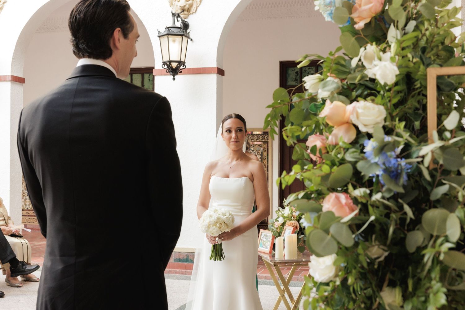 A couple walking together through a beautifully decorated wedding archway adorned with white flowers and greenery.