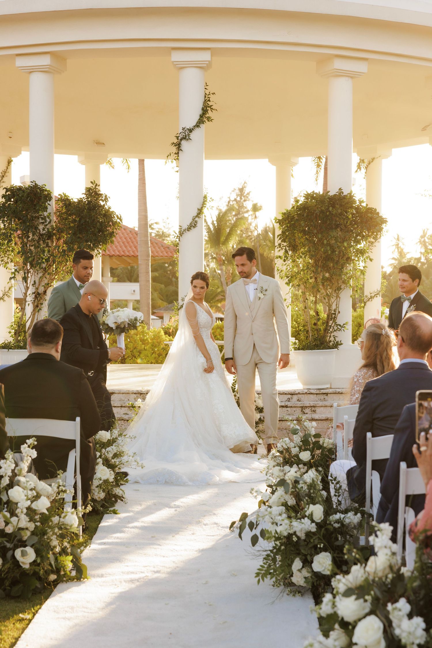 A wedding ceremony setup in an elegant covered terrace with white floral arrangements and columns at sunset.