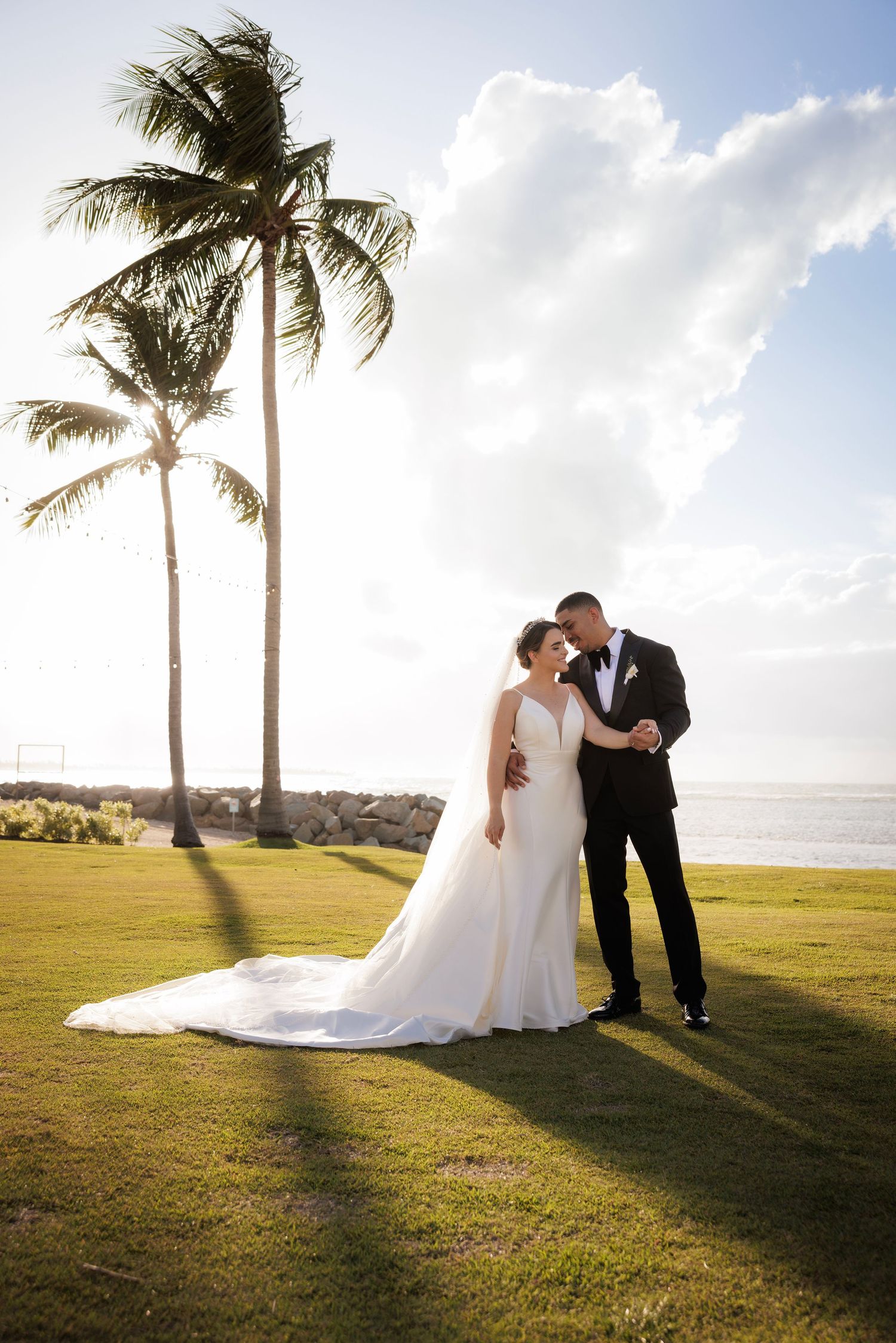 A couple shares romantic moments at sunset on a tropical beach with palm trees and rolling hills in the background.