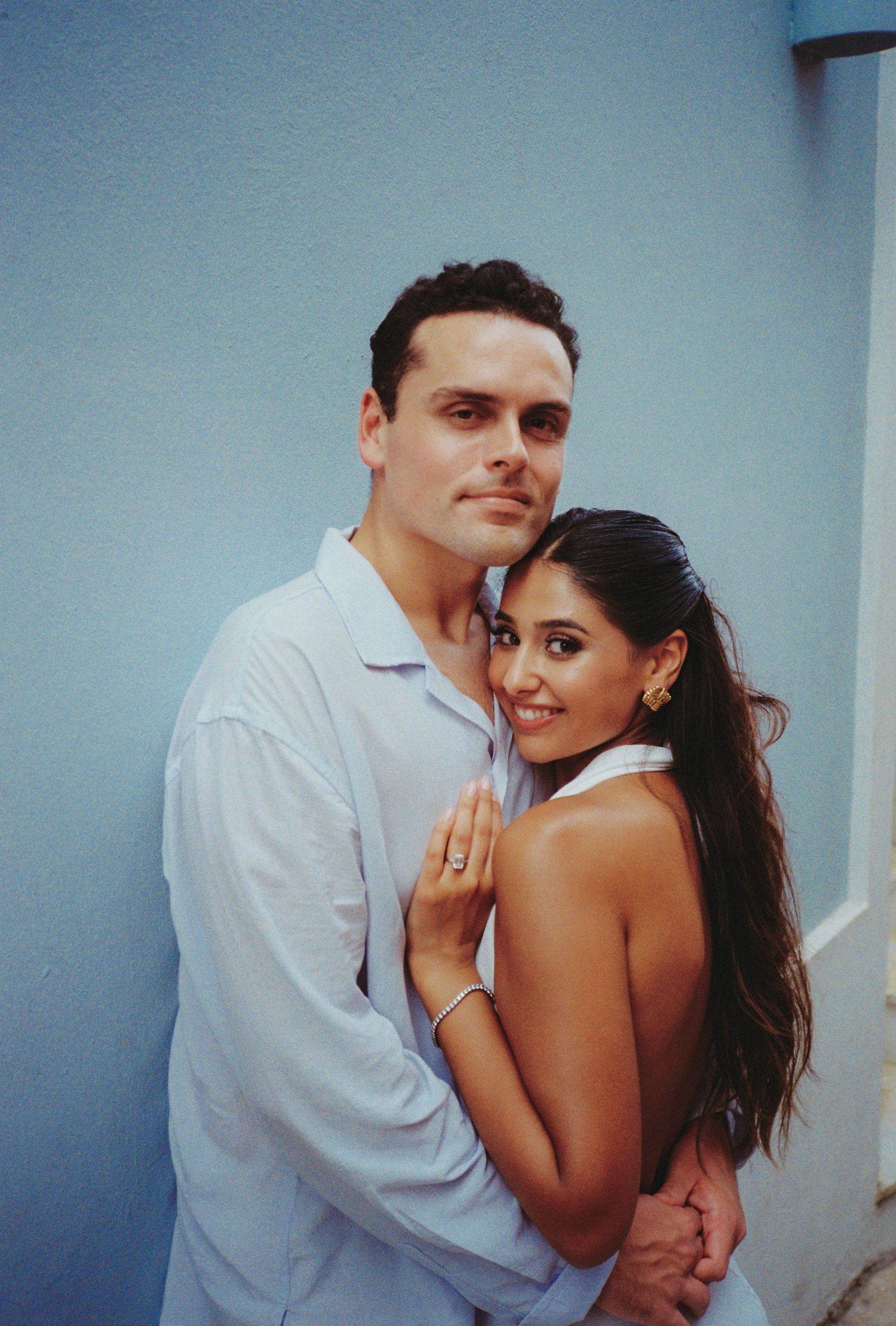 Couple embracing against blue wall, woman in backless white dress with ring, man in white shirt, engagement photo.