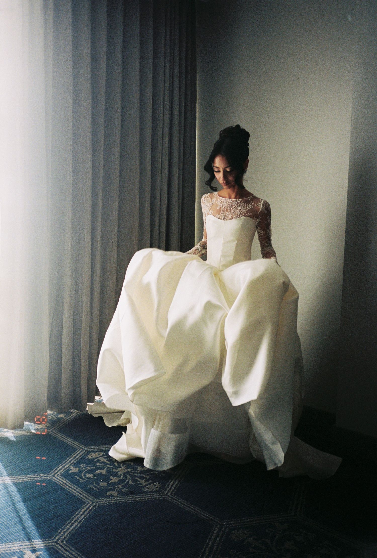 Bride in elegant white lace wedding gown holding full skirt near window in soft natural light.