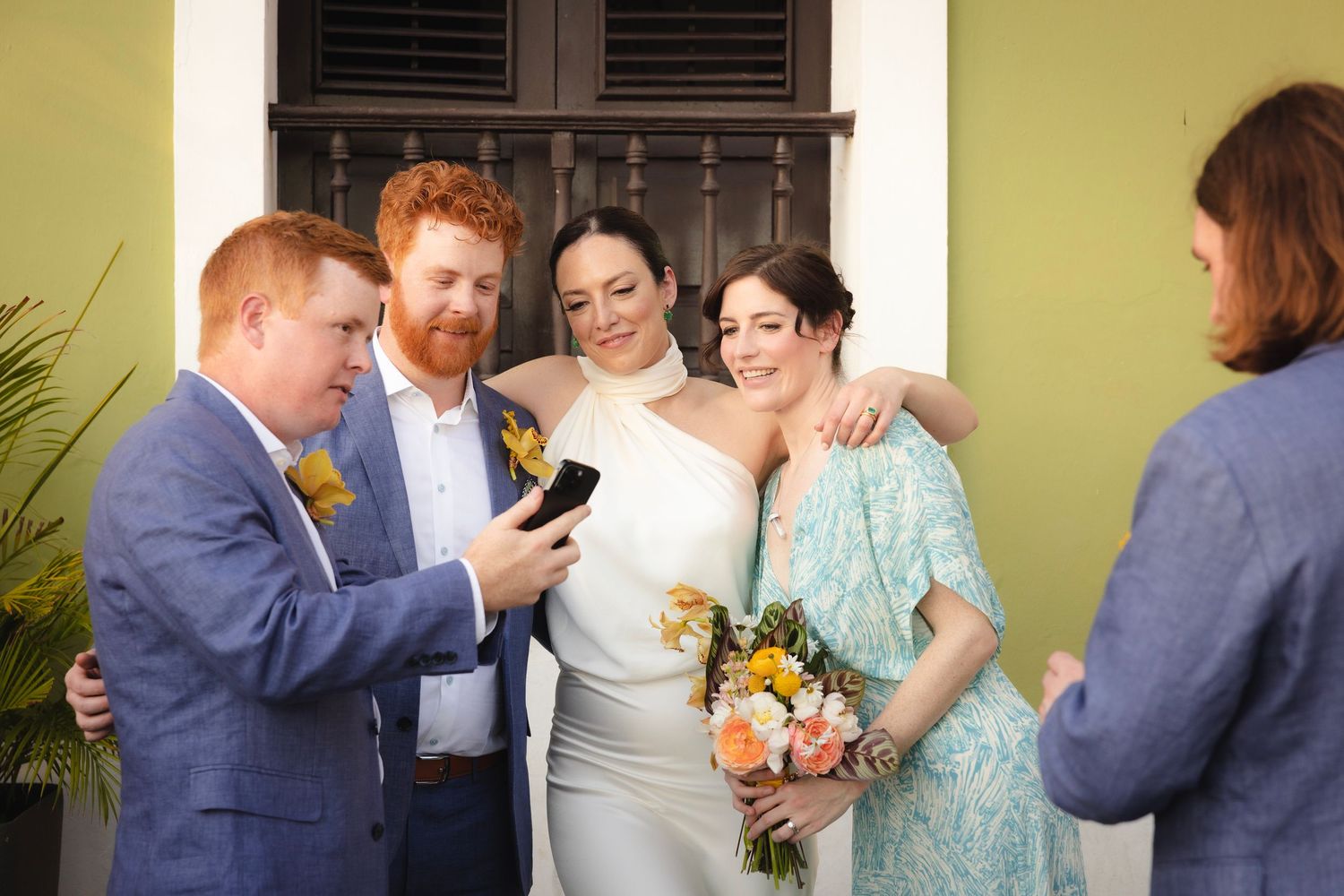 Wedding party members sharing candid moments against yellow wall.