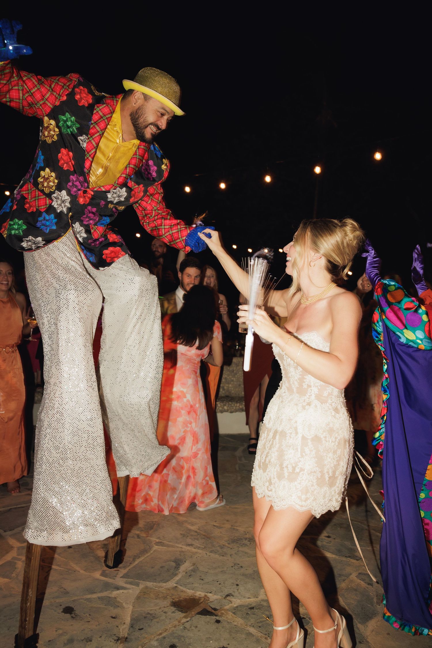 Lively party scene on a beach at night with people dancing under string lights in colorful festive outfits.
