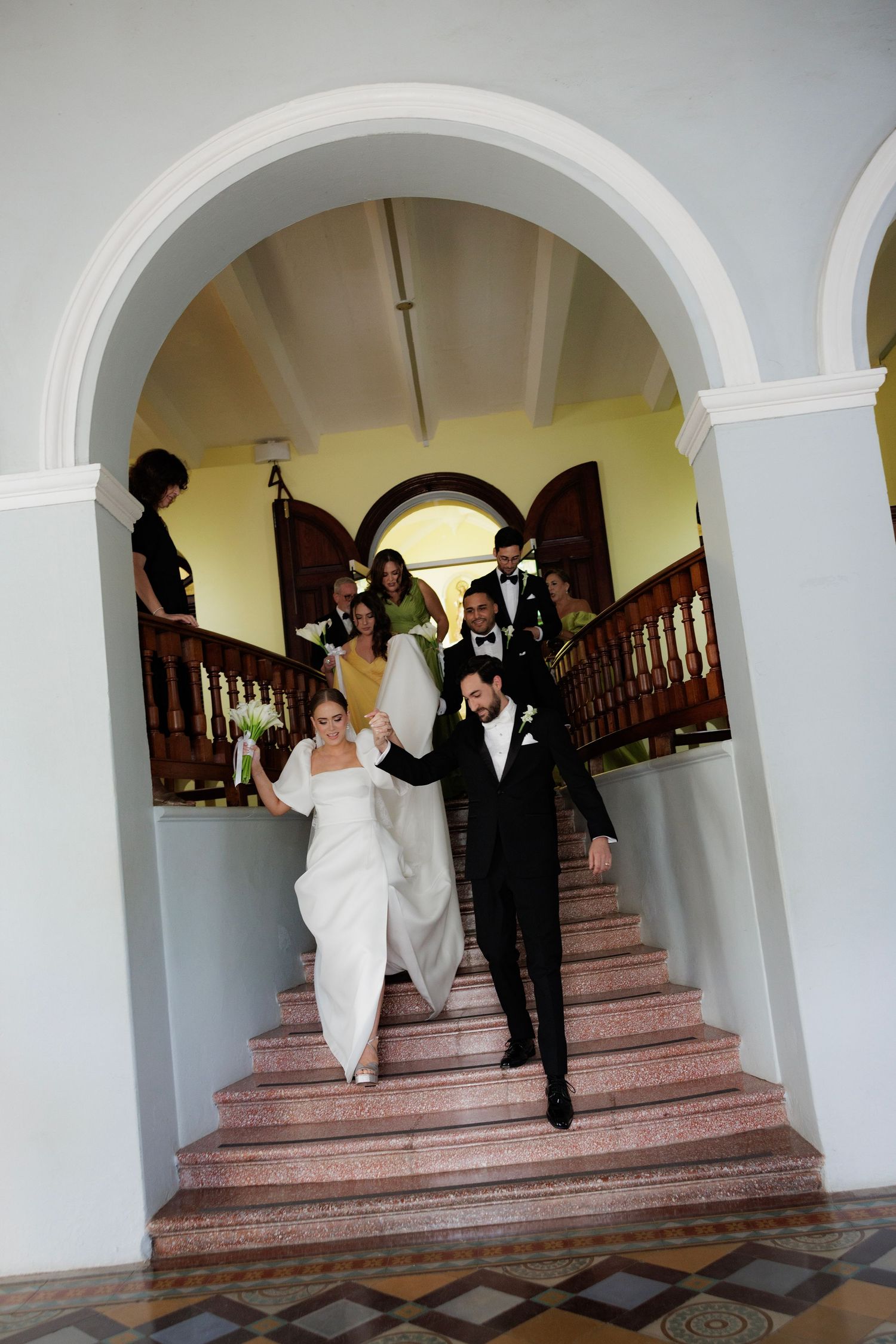 A newlywed couple descends a grand staircase under an arched walkway after their ceremony.