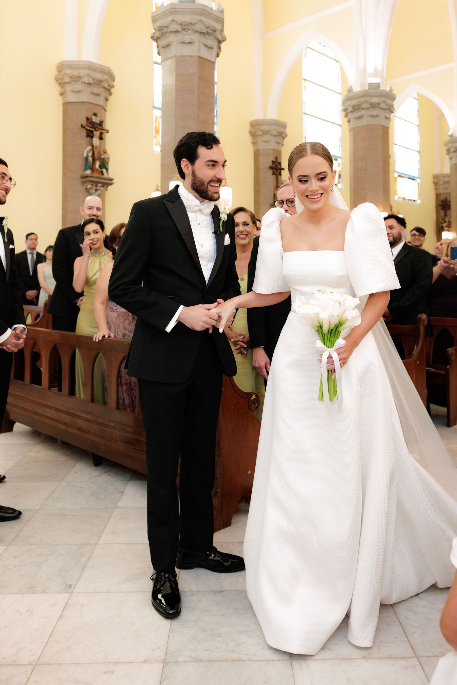A wedding couple walks down the aisle of a church with ornate stone columns and yellow walls.