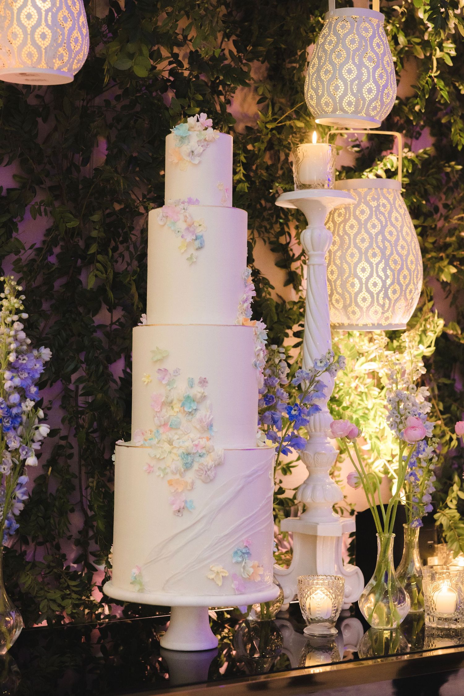 Elegant white tiered wedding cake decorated with pastel blue and pink flowers, illuminated by glowing lanterns and candlelight.