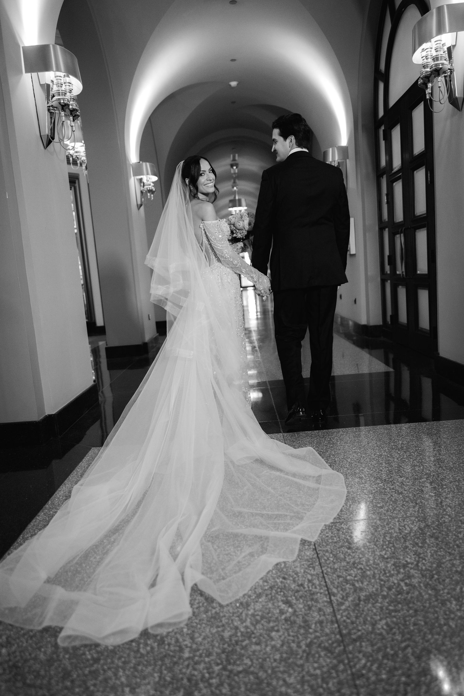 Black and white photo of a bride and groom in an arched hallway with dramatic lighting.