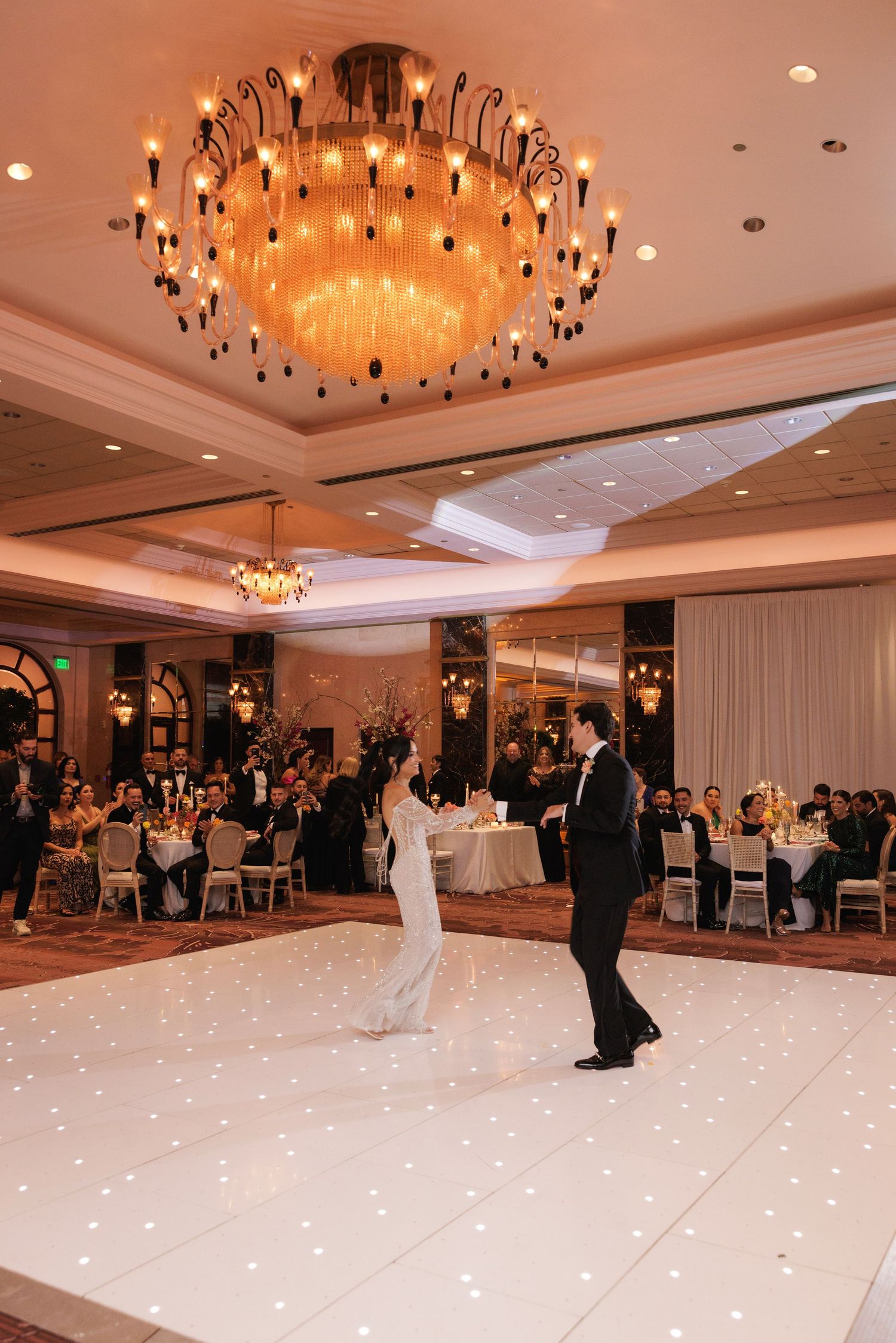 A formal dance taking place in an opulent ballroom with a glowing chandelier and white illuminated dance floor.