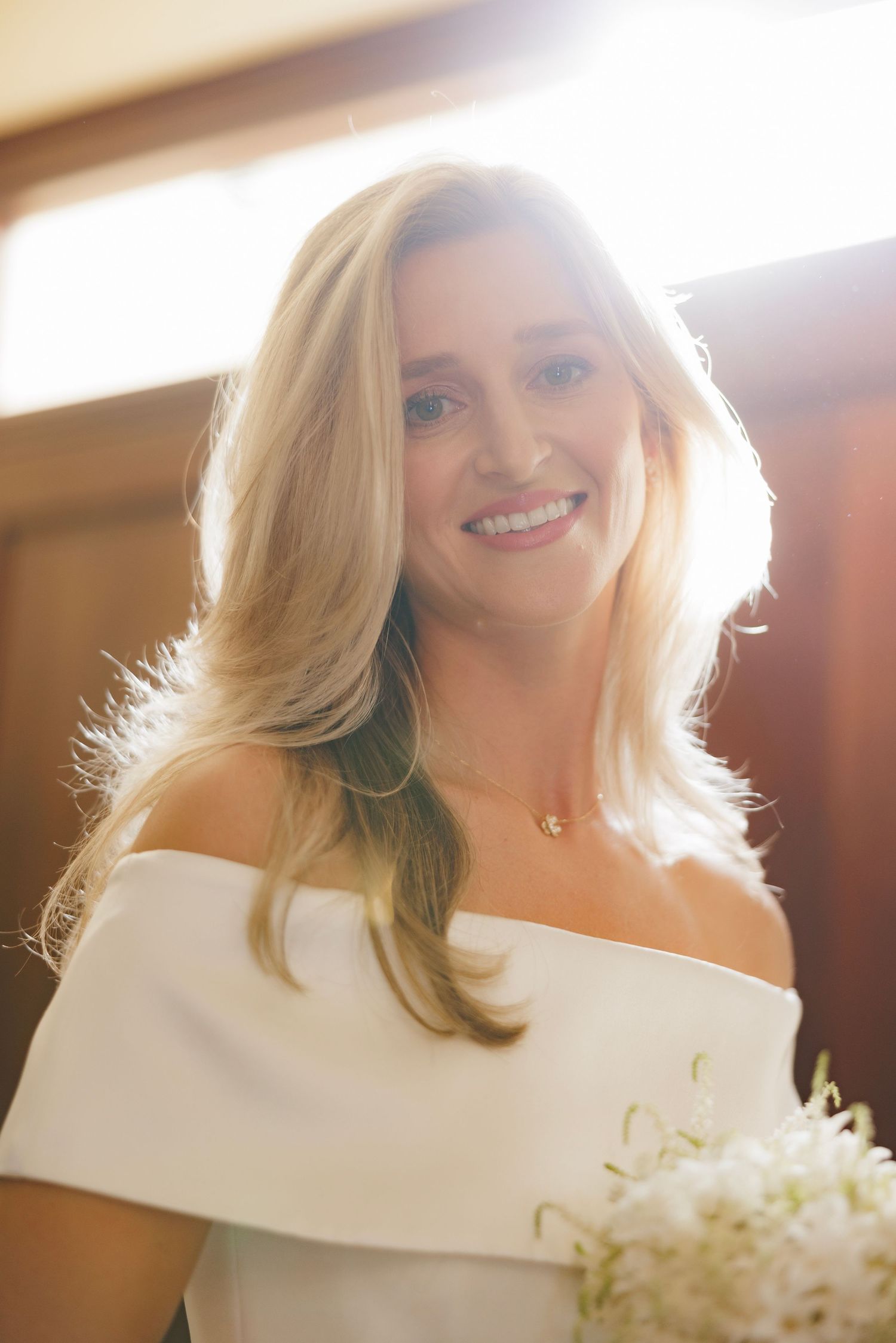 Sunlit portrait of someone in an off-shoulder white dress with blonde hair and a bright smile holding white flowers.