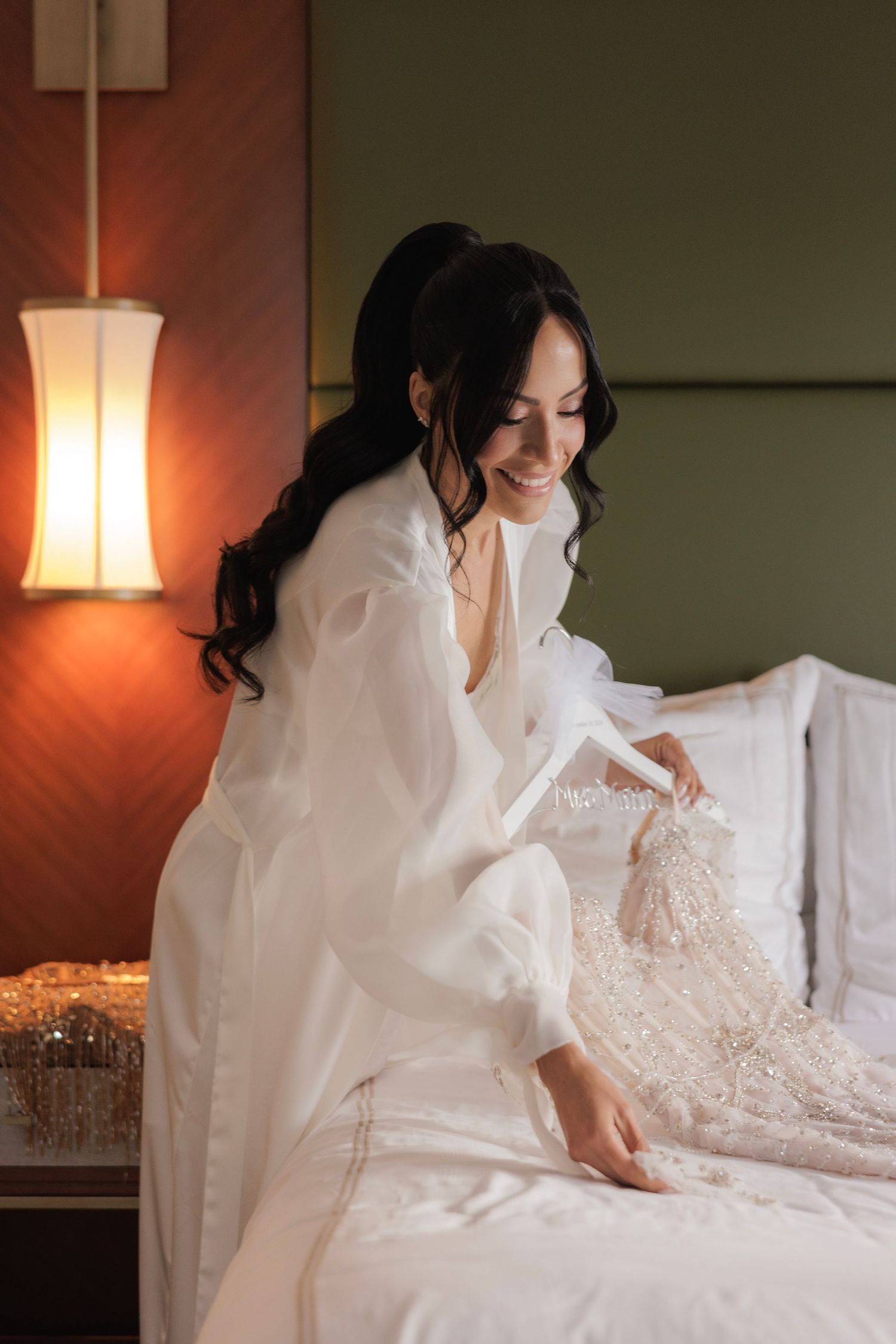 White lace wedding dress laid out on bed with warm lighting from bedside lamp.