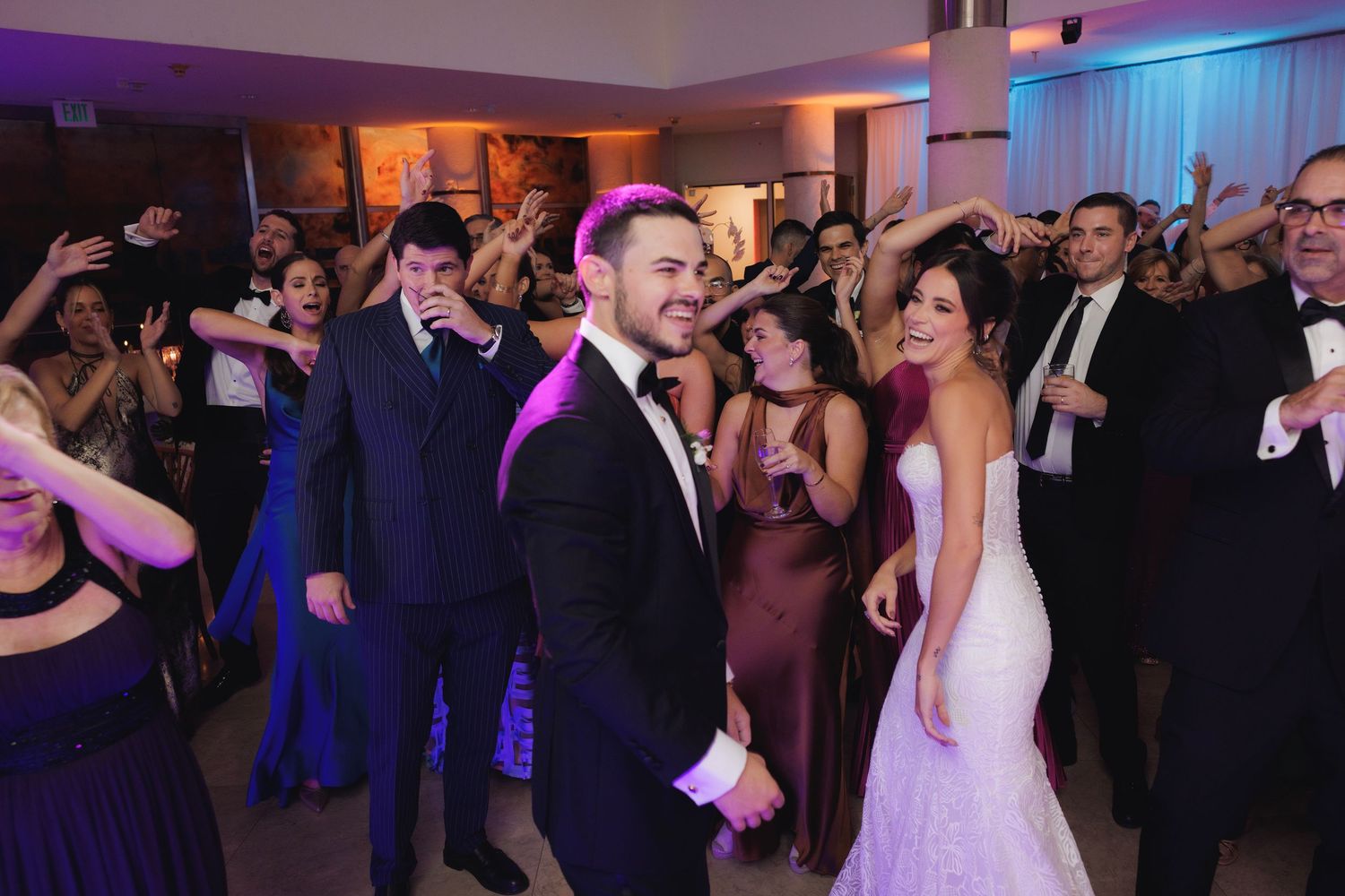 Wedding guests dance and celebrate on a vibrant dance floor illuminated with purple and warm lighting.