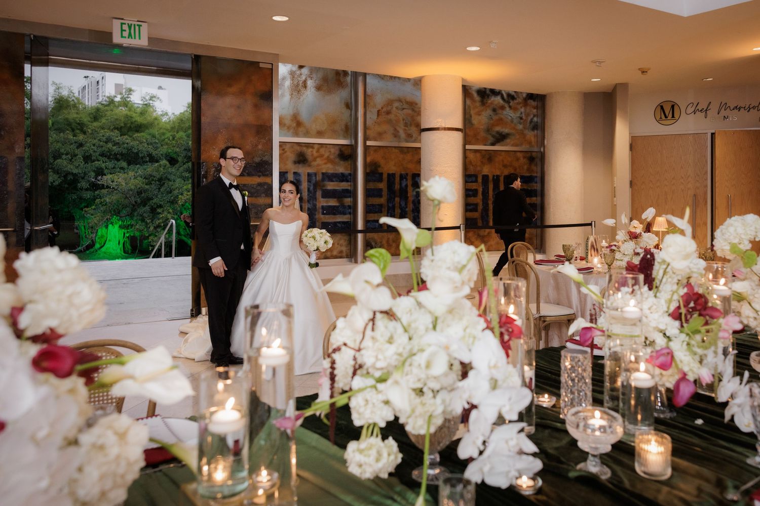 Elegant wedding reception tables decorated with white orchids, pink roses, and glowing candles in a modern venue.