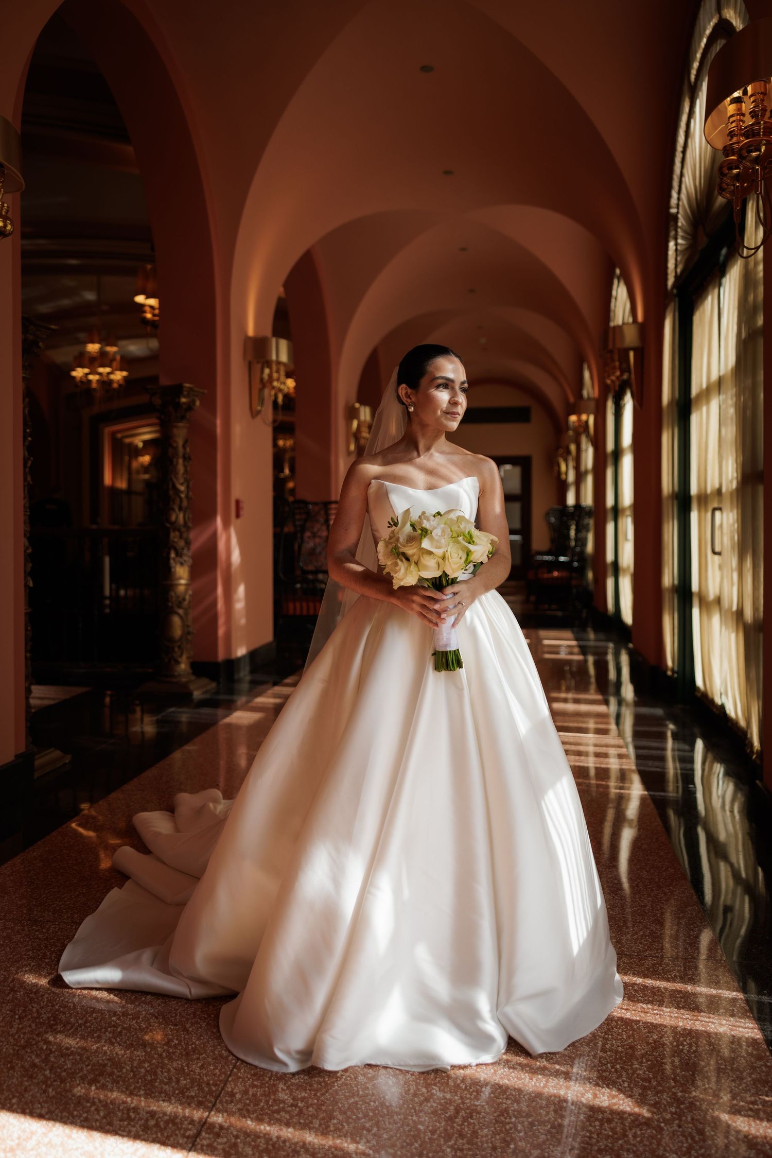A bride in an elegant white ballgown poses in a grand hallway with arched ceilings and natural lighting.