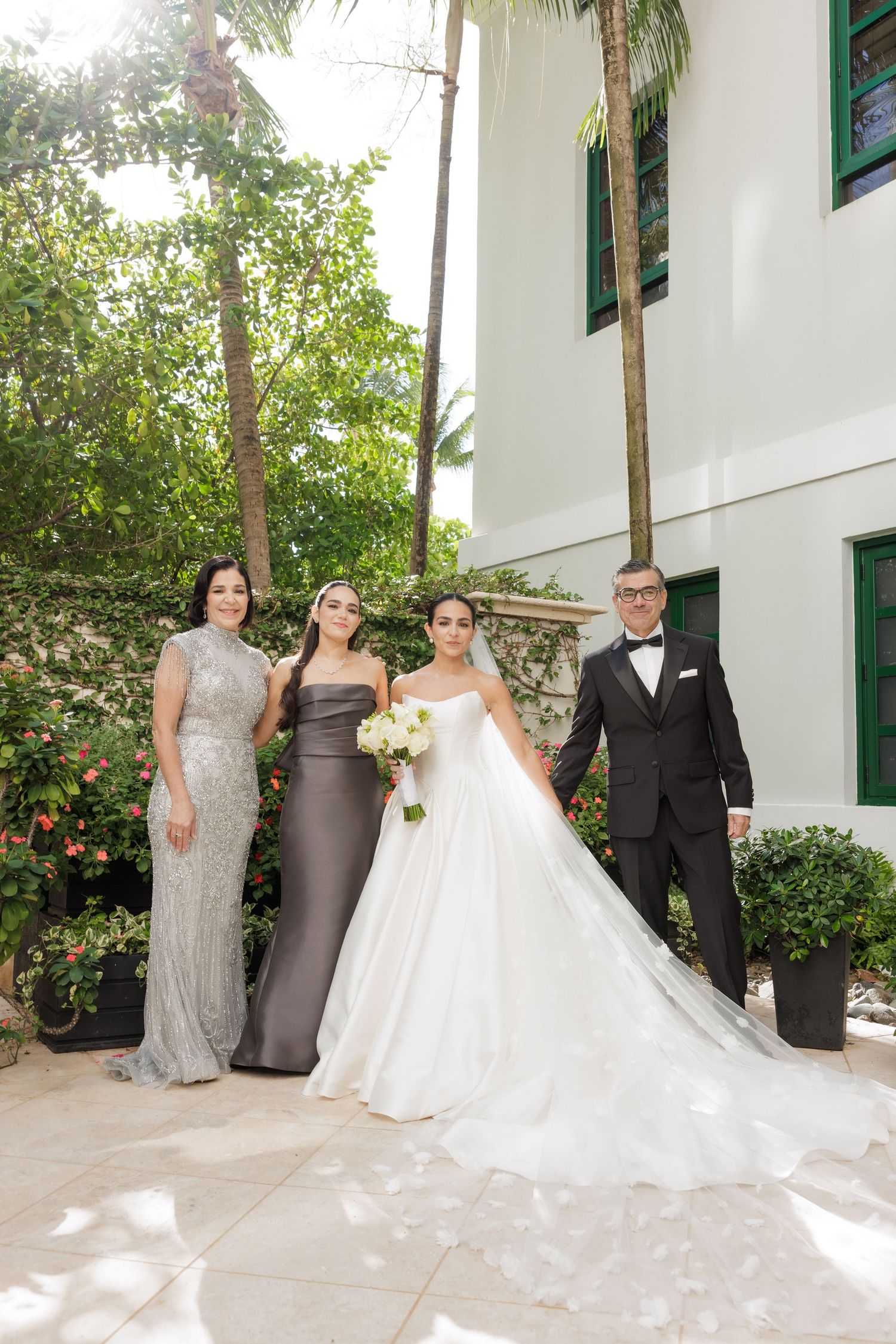 Wedding party poses outdoors against white building with tropical palm trees and greenery.