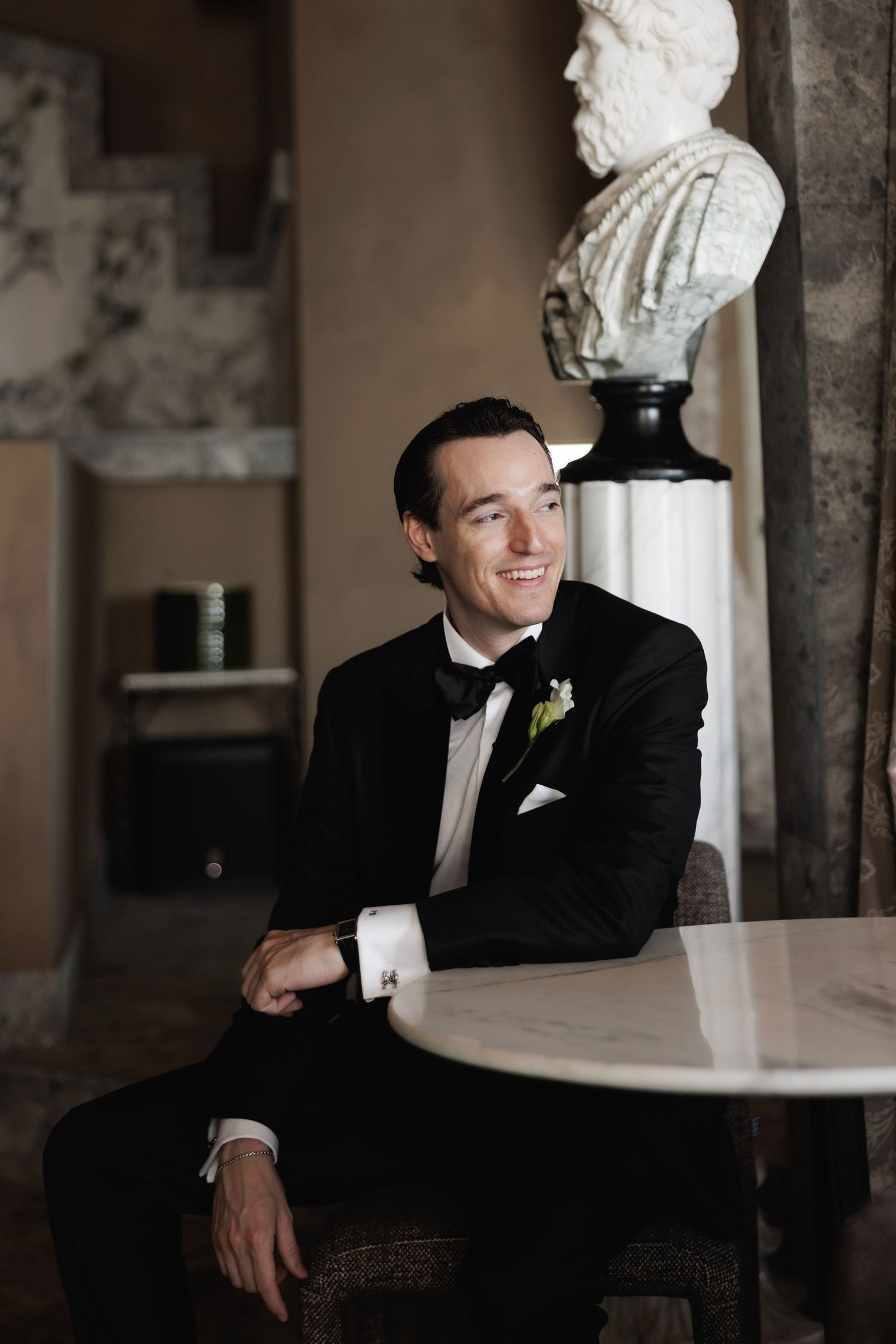 Groom in classic black tuxedo sitting at a table in an elegant venue with white floral arrangements.