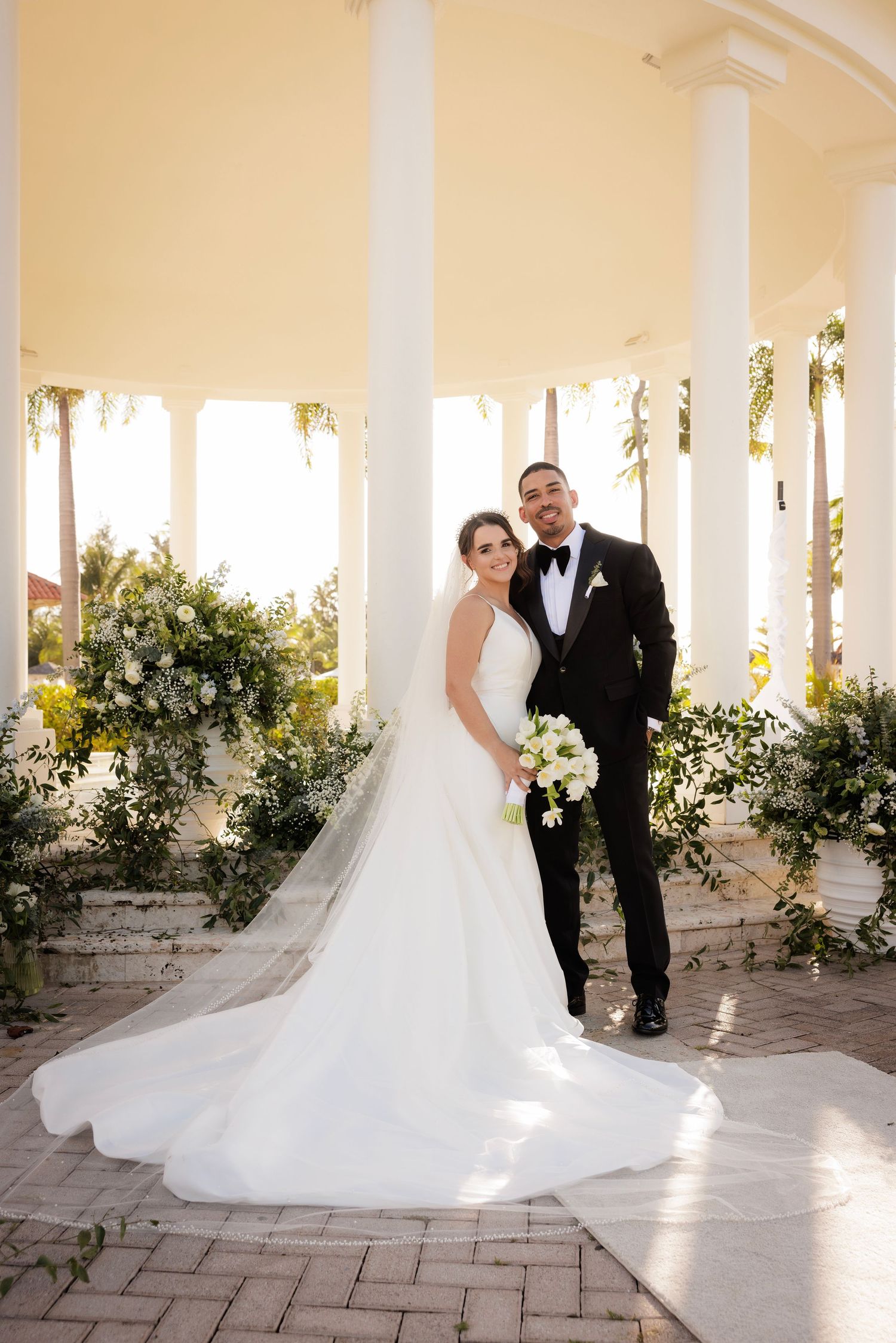 Wedding couple poses elegantly under white columns at an outdoor venue with lush floral arrangements.