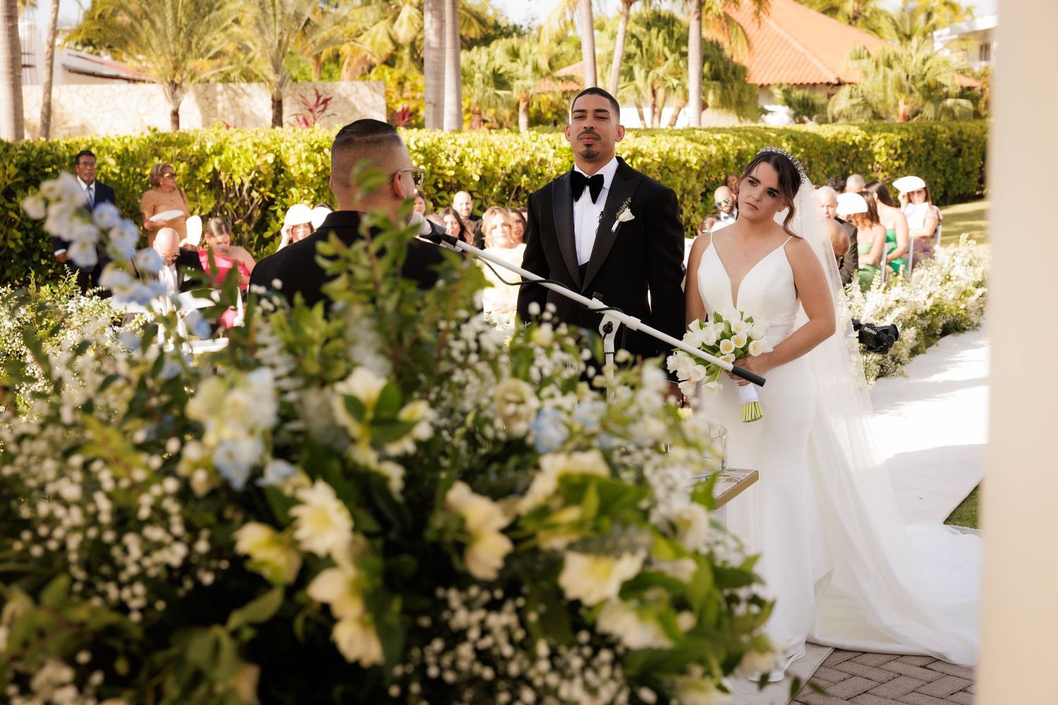 Couple in wedding attire strolls together under a canopy of lush palm trees and greenery at tropical venue.