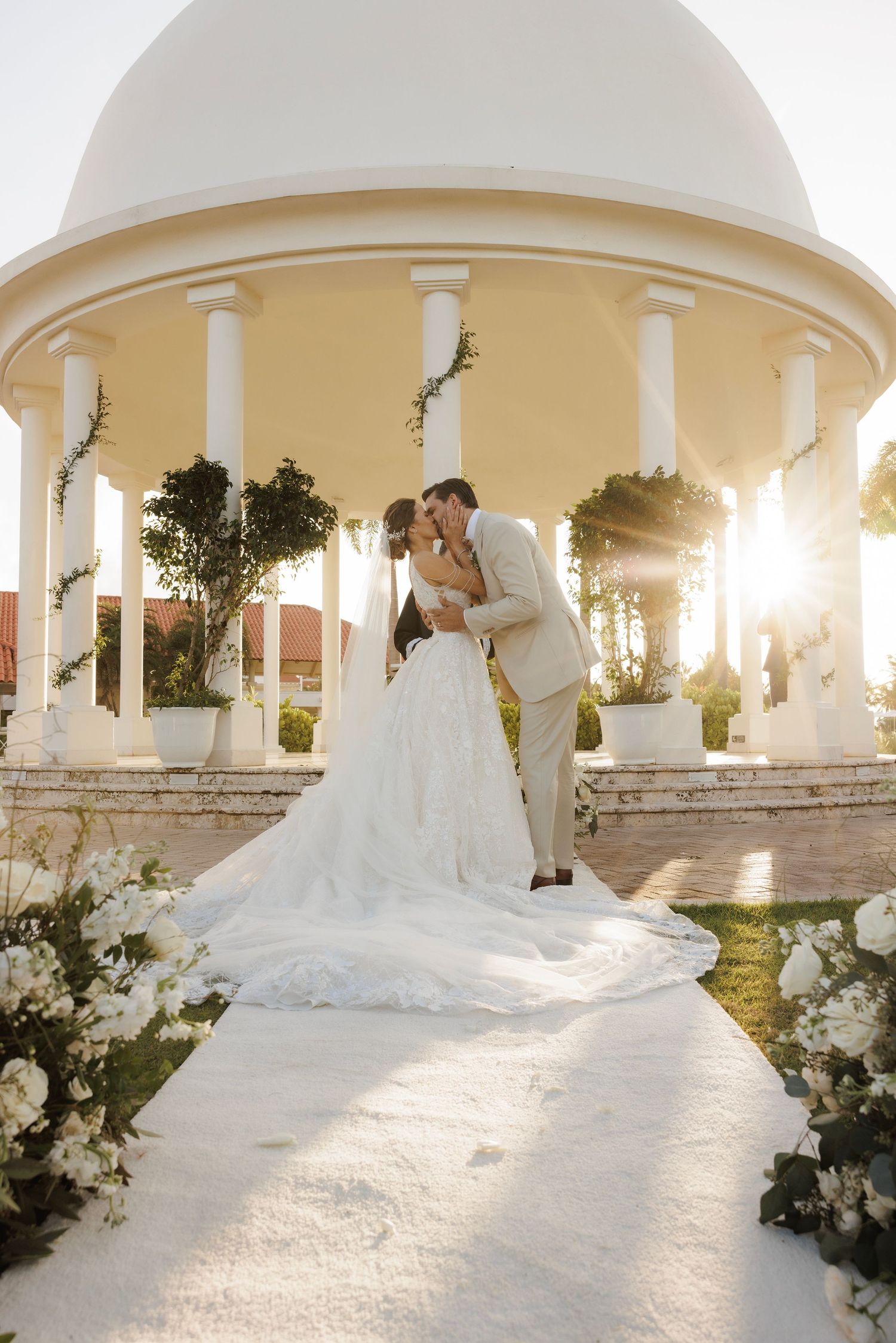 A stunning white wedding dress with a dramatic cathedral train photographed in front of a columned gazebo at sunset.