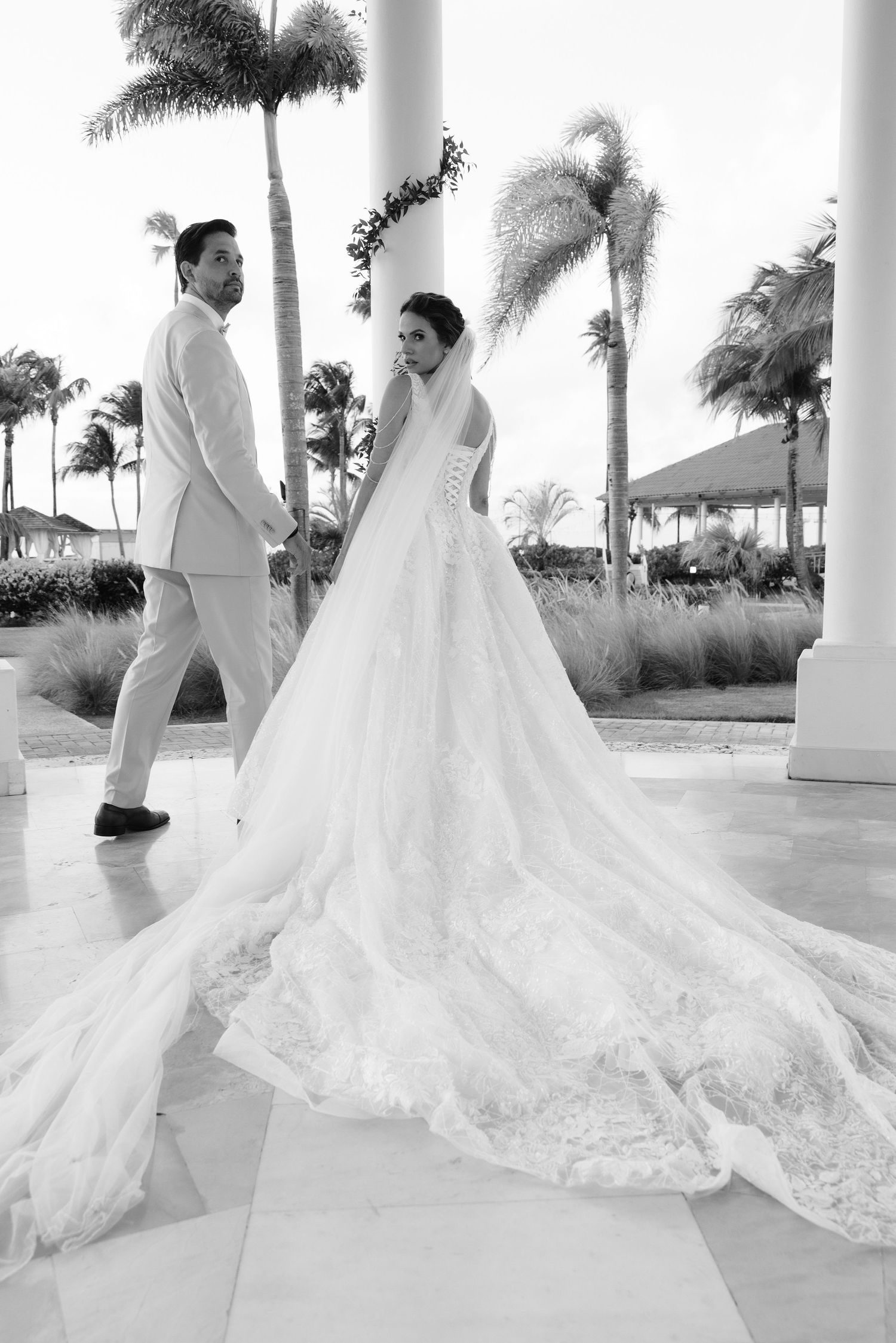Black and white wedding photo showing a couple in a tropical resort setting with palm trees and elegant white columns.