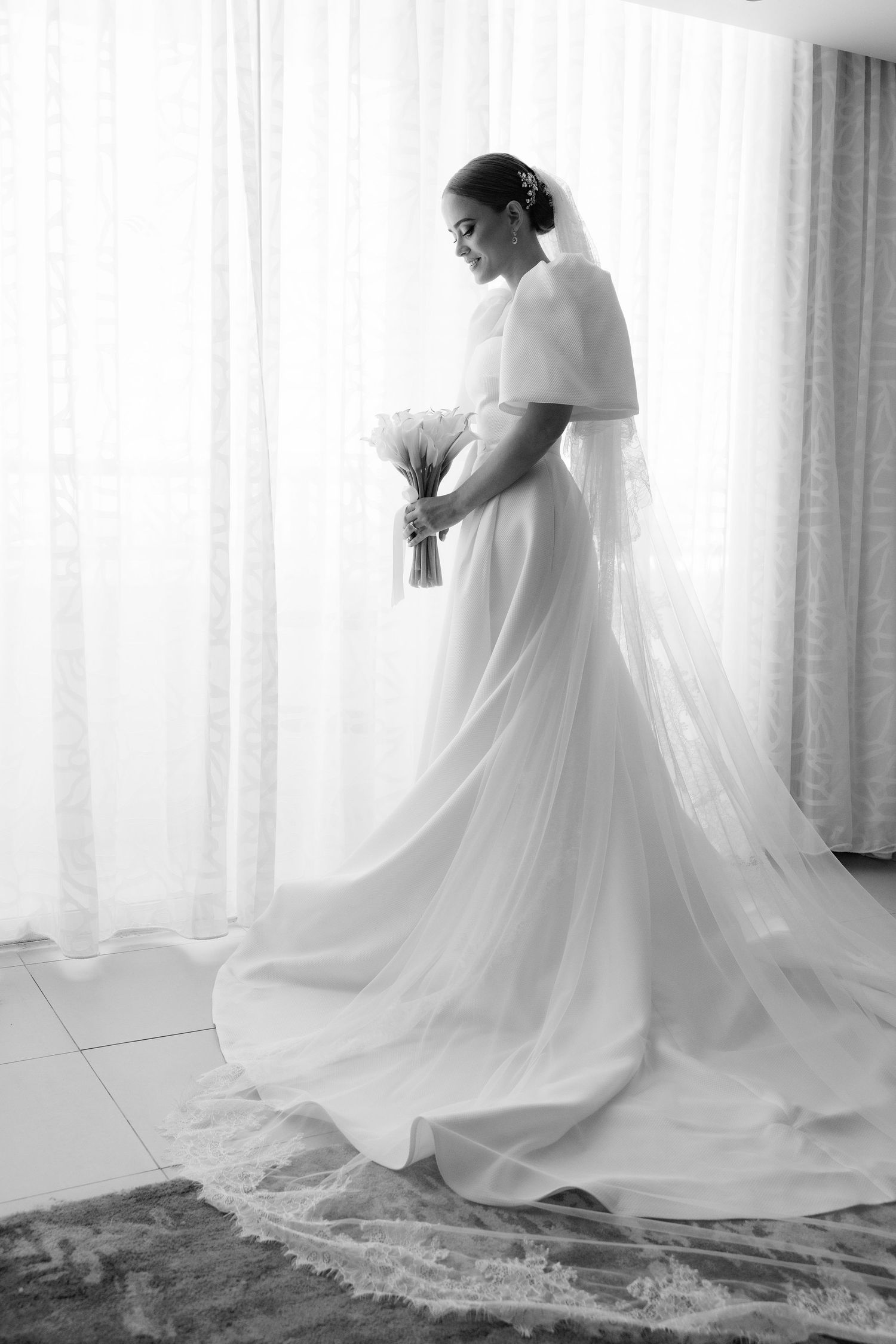 Elegant bride in flowing white gown posing by window in soft natural light.