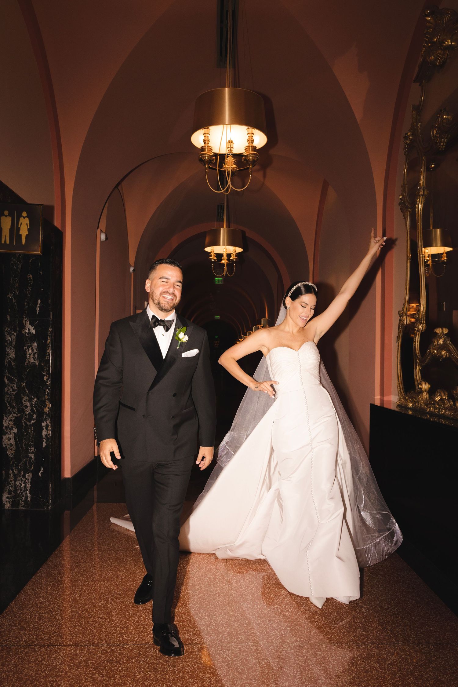 A couple dancing and having fun at a wedding reception in an elegant venue with arched ceilings and chandeliers.