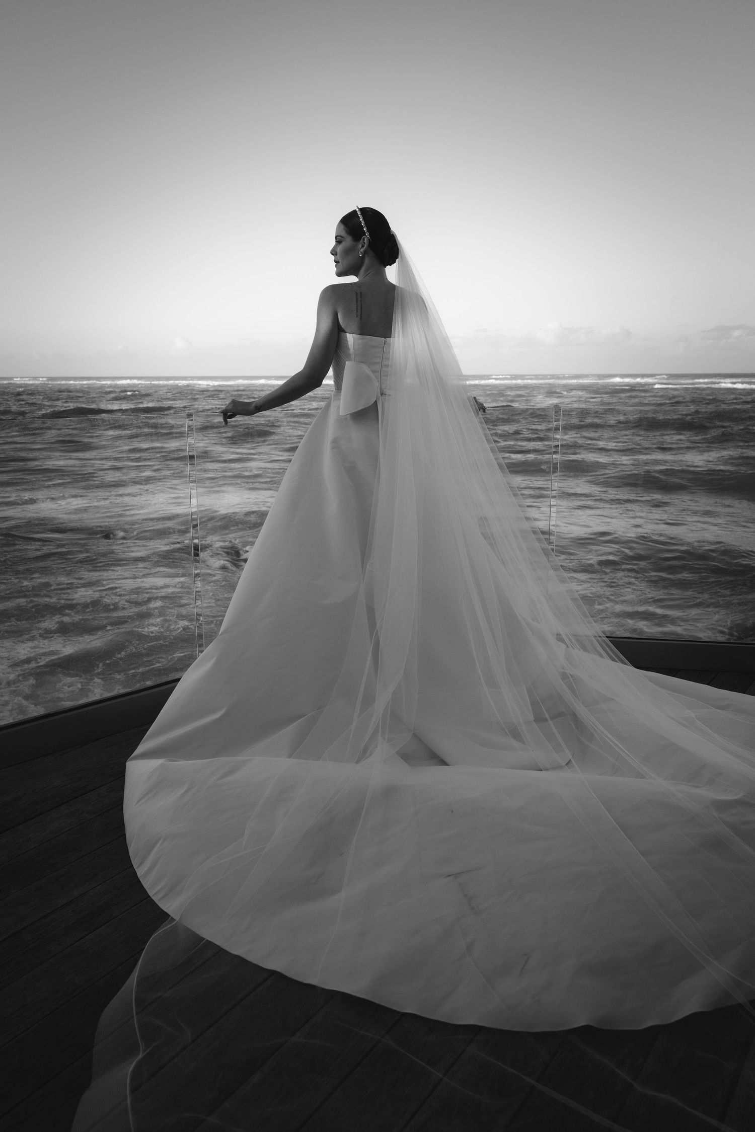 A bride in a flowing white dress and veil stands silhouetted against the ocean at sunset in black and white photography.