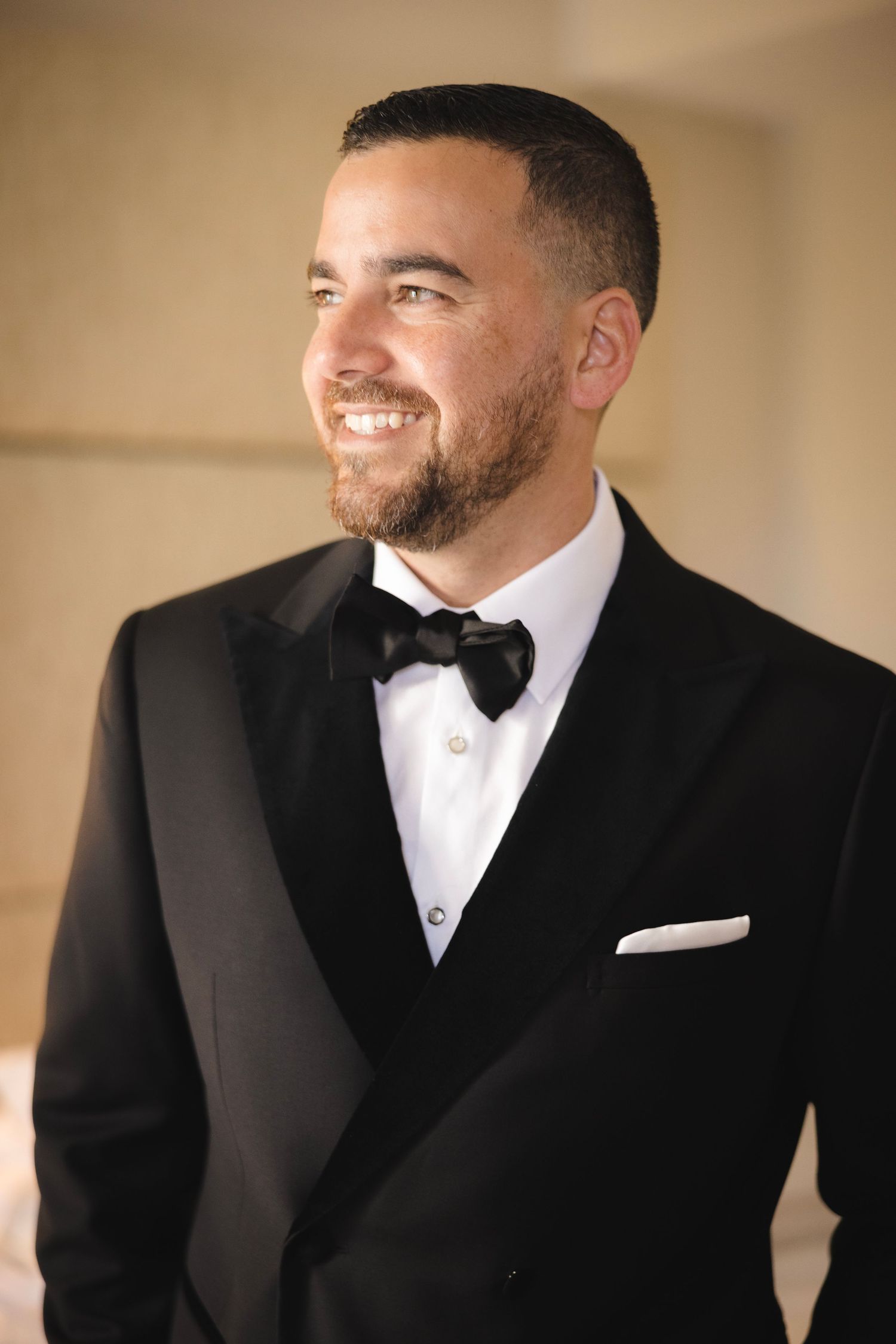 A groom in a black tuxedo and white bowtie smiles while looking to the side in a formal portrait series.
