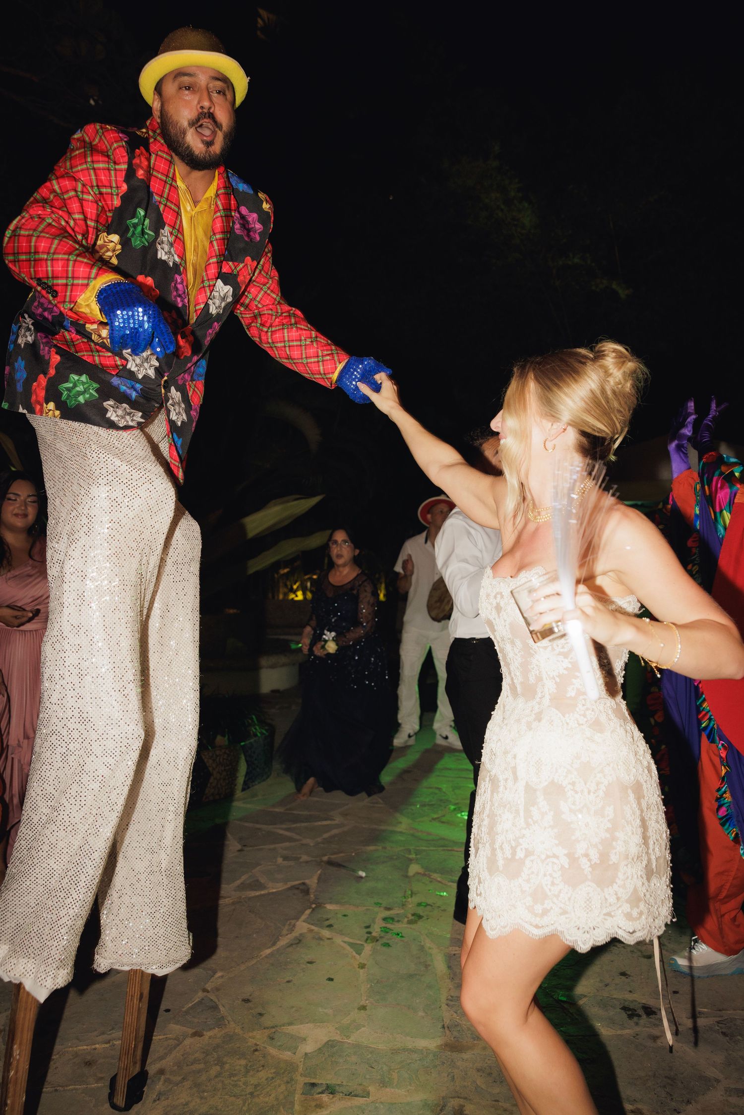 A festive party scene shows two dancers, with one wearing a vibrant patterned jacket and stilts, and a white dress on the other.