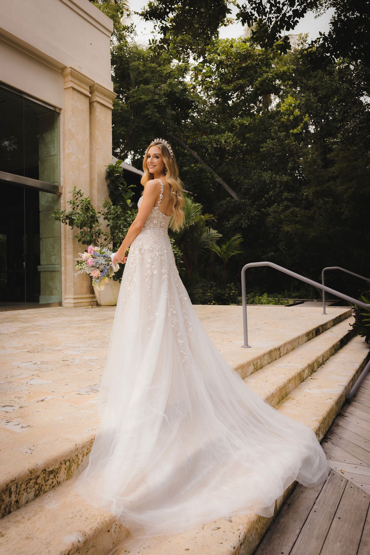 A flowing white wedding dress with a dramatic train cascades down elegant stone steps in an outdoor garden setting.