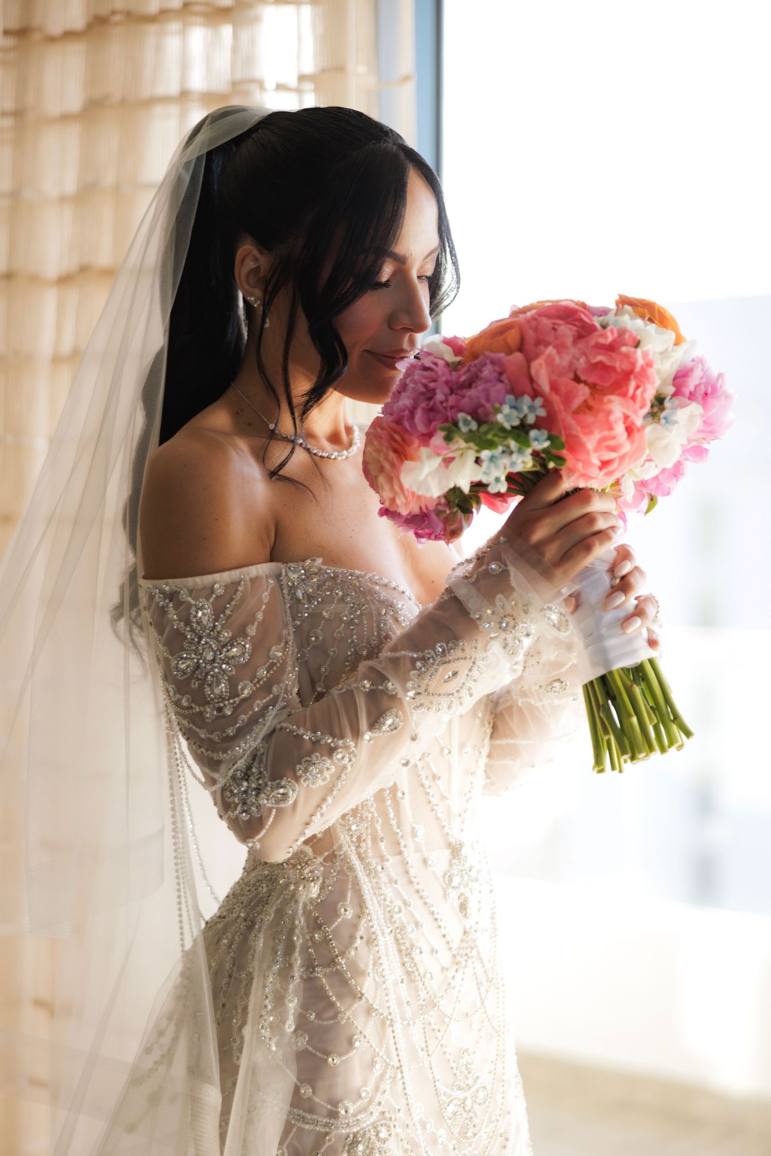 Bride in lace gown admires pink and coral wedding bouquet near window.