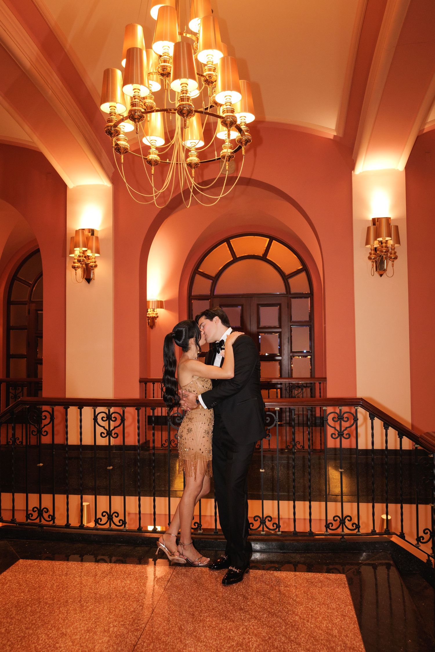 A romantic moment under a grand chandelier in an elegant ballroom with arched windows and blush-toned lighting.