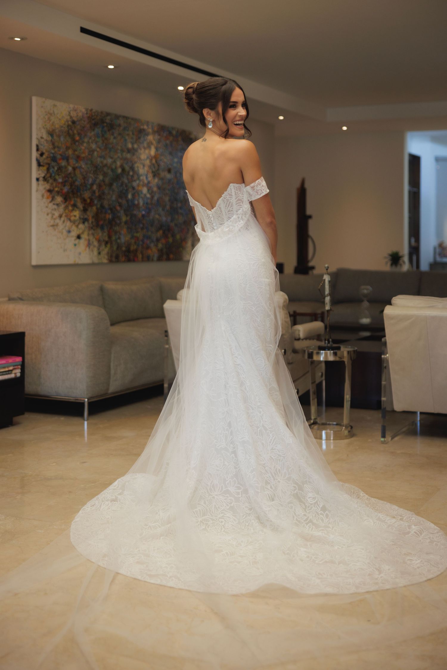 Back view of bride in off-shoulder lace wedding gown with long train standing in modern living room.