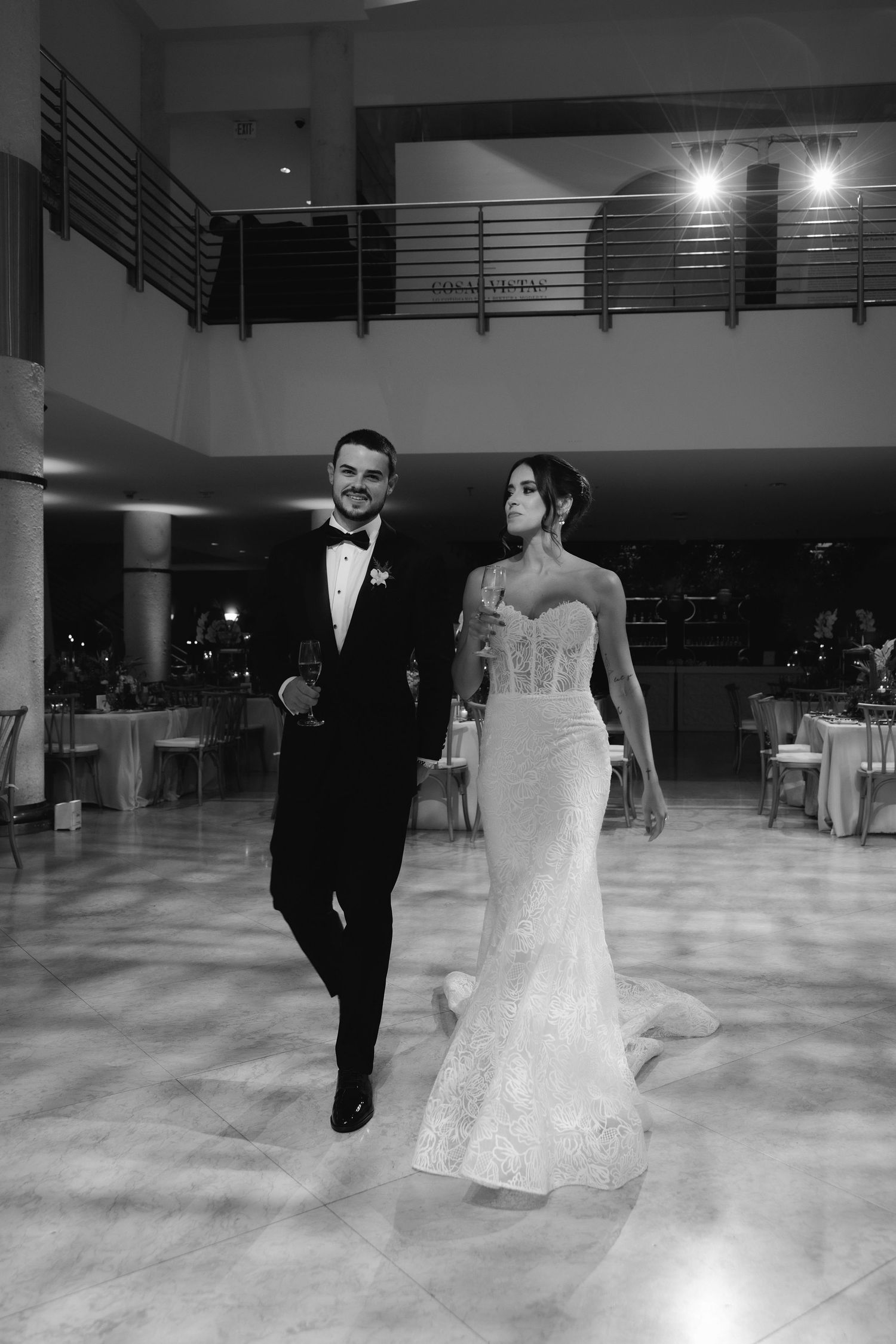 A bride in a fitted lace gown and groom in classic tuxedo walk together on their wedding day in a modern reception venue.