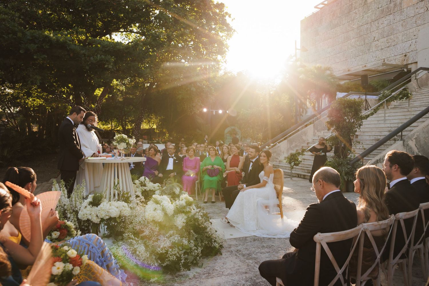 Sun flares illuminate an outdoor wedding reception area with stone walls and greenery at golden hour.