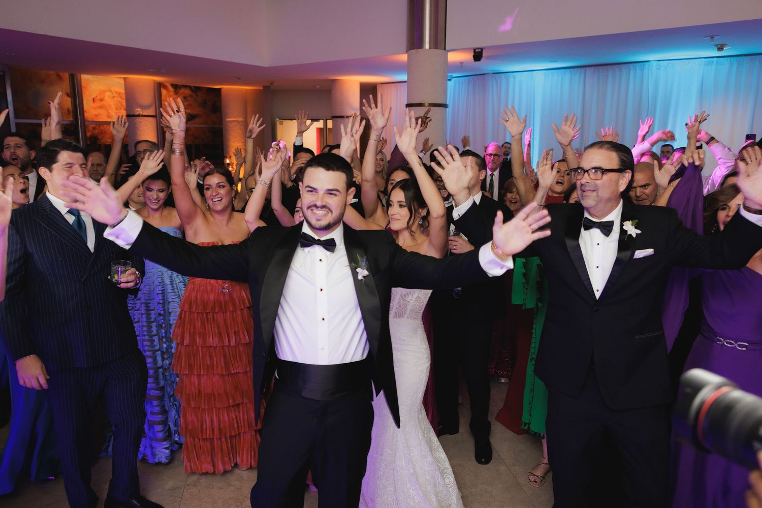 Wedding reception guests dancing and celebrating on a colorfully lit dance floor in formal attire.