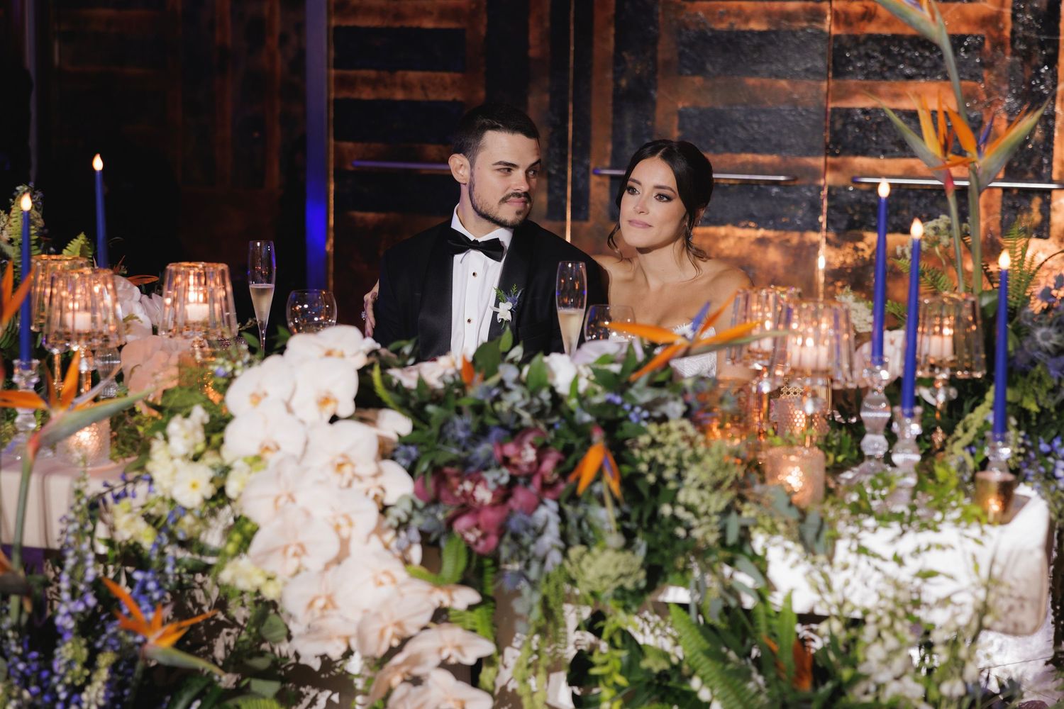 Elegant wedding reception table decorated with lush floral arrangements, glowing candles and a tiered white cake.