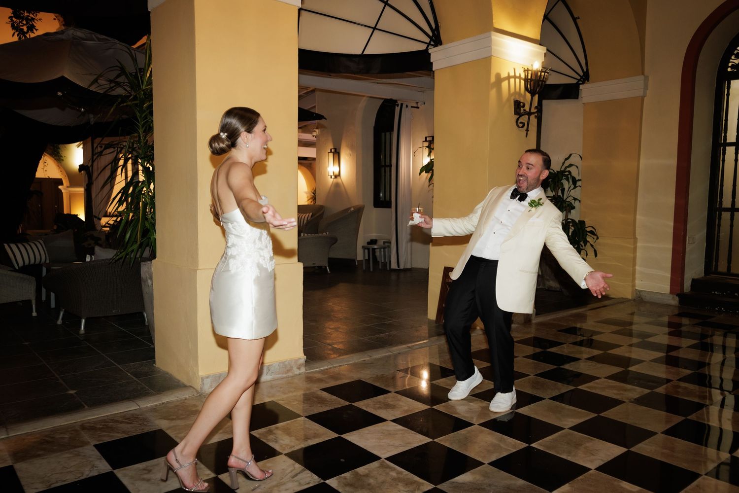 Two people dancing enthusiastically on a black and white checkered floor in an elegant venue with arched windows and warm lighting.