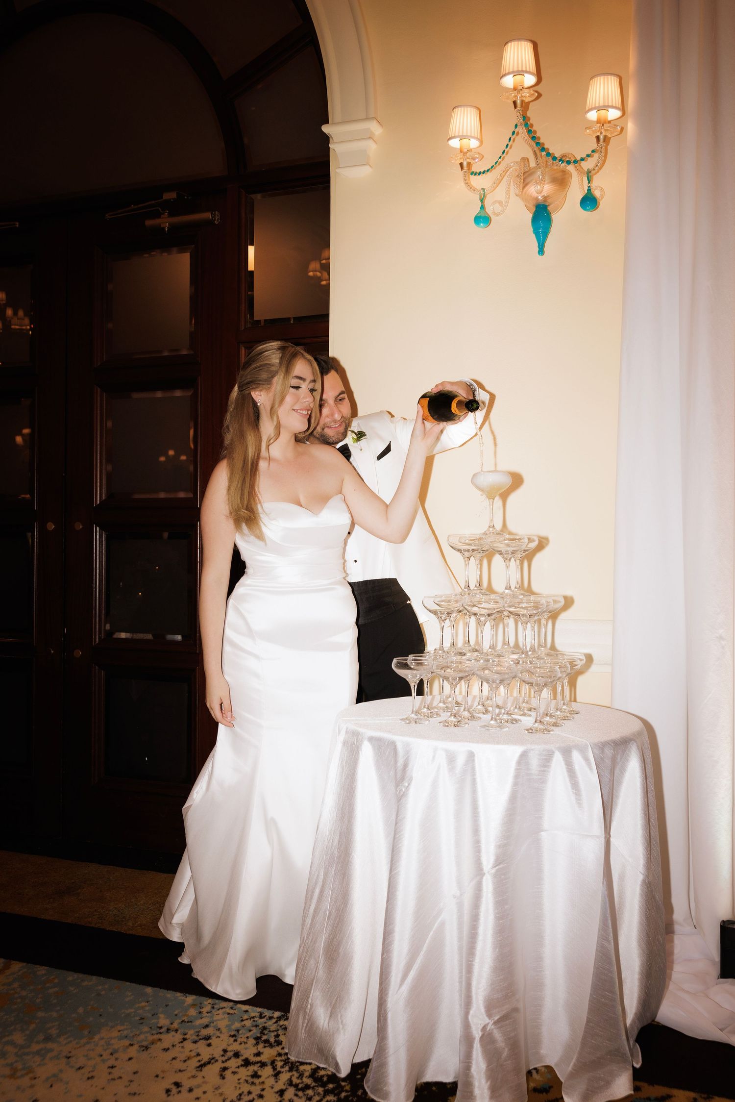 Wedding reception venue with champagne tower and elegant white table settings illuminated by warm lighting.