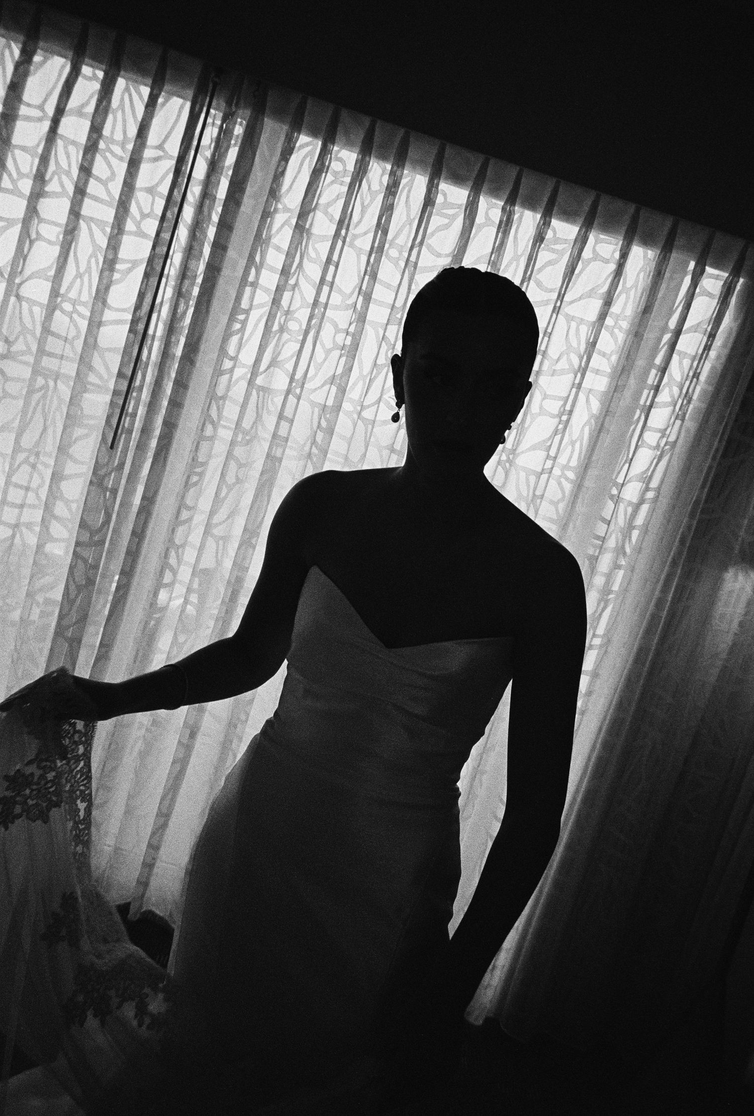 Dramatic silhouette of bride in strapless wedding gown standing before sheer curtained window.