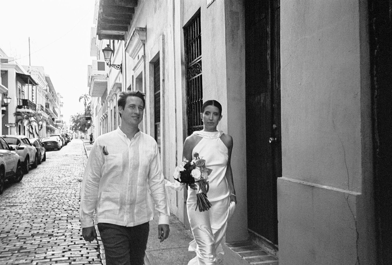 Black and white photo of a couple walking on a cobblestone street, bride in white dress holding bouquet, groom in white shirt.