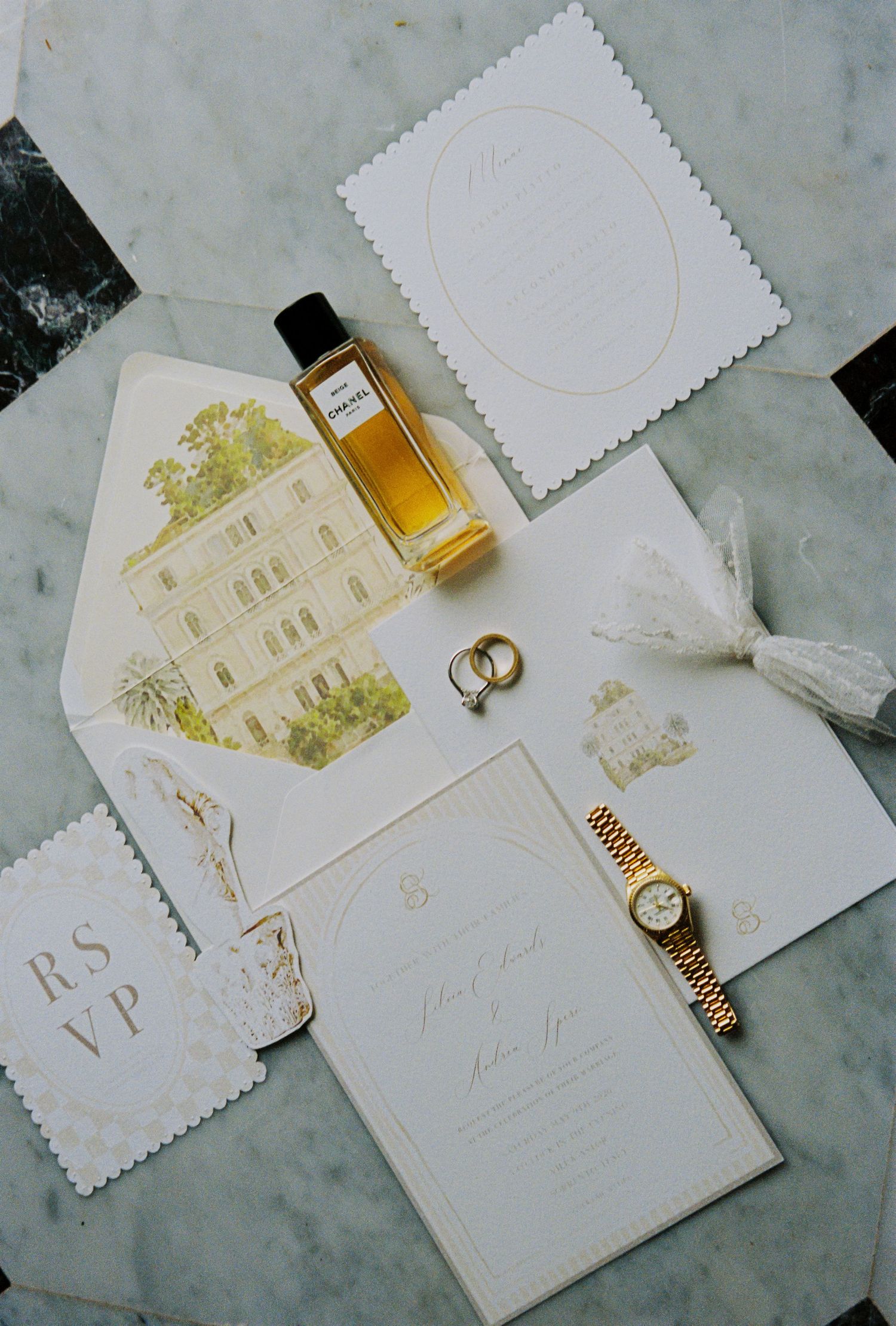 Elegant wedding invitation suite with gold accents, RSVP card, perfume bottle, rings, and a Rolex watch on marble.