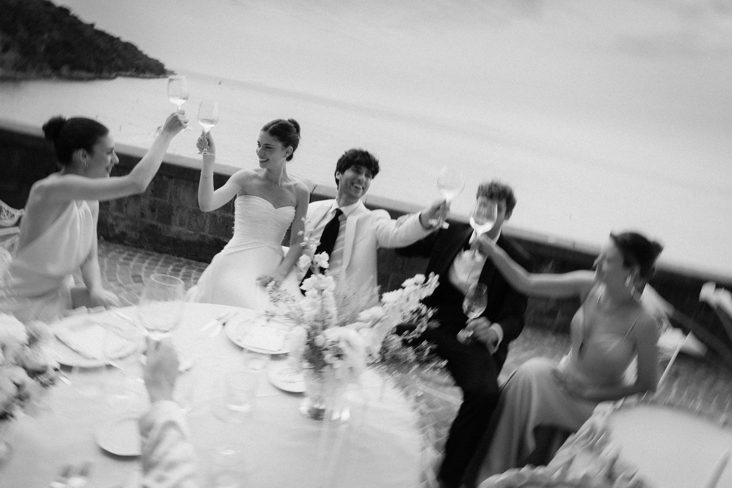 Black and white wedding reception toast with bride, groom, and guests raising glasses outdoors by the sea.