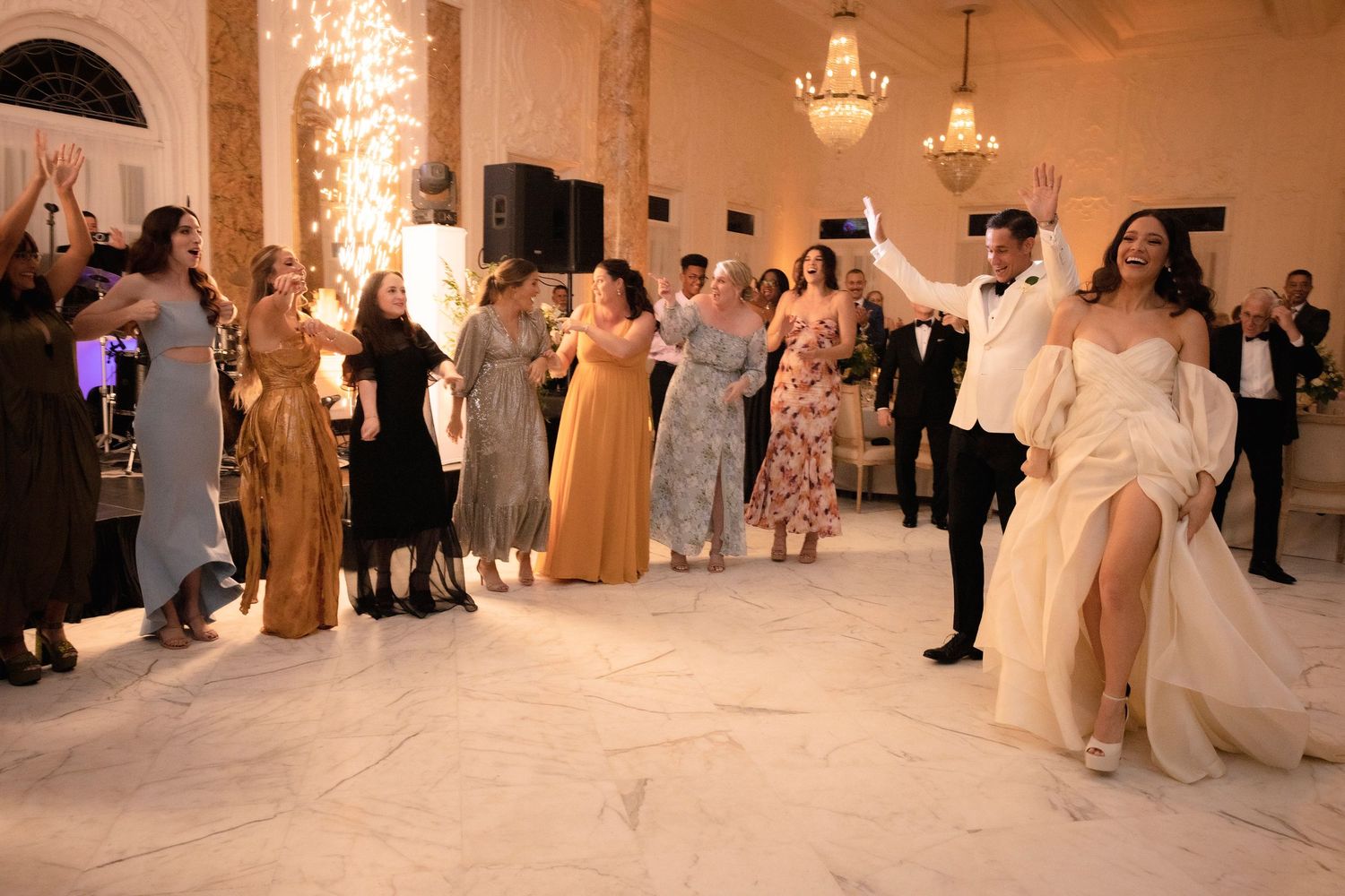 Wedding guests dancing and celebrating at an elegant ballroom reception with marble columns.